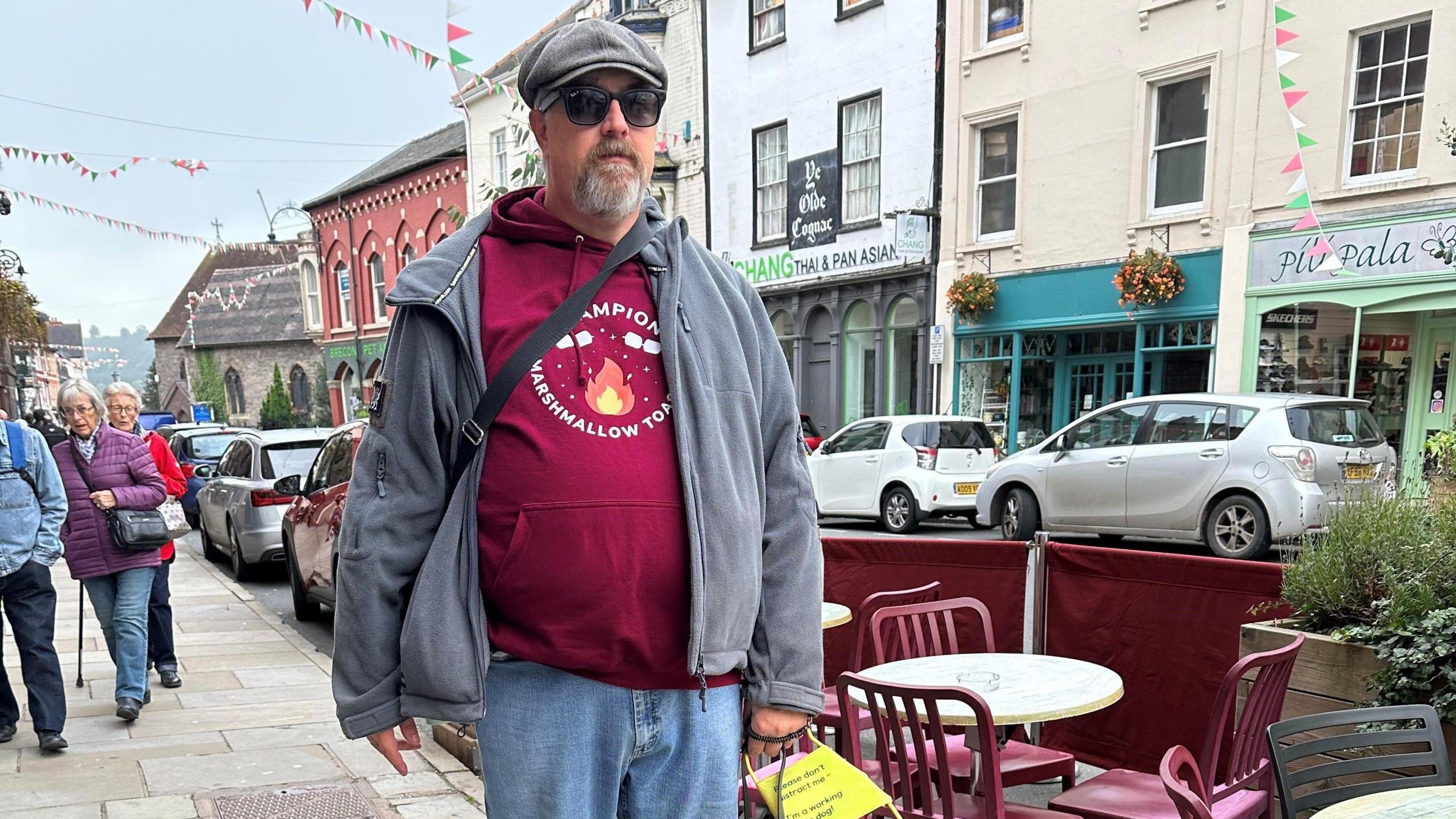 A man standing on a pavement with pedestrians behind him and a restaurant outside seating between him and the road. He has a open grey hoodie, dark glasses and a flat cap on. He has a grey goatee style beard. 