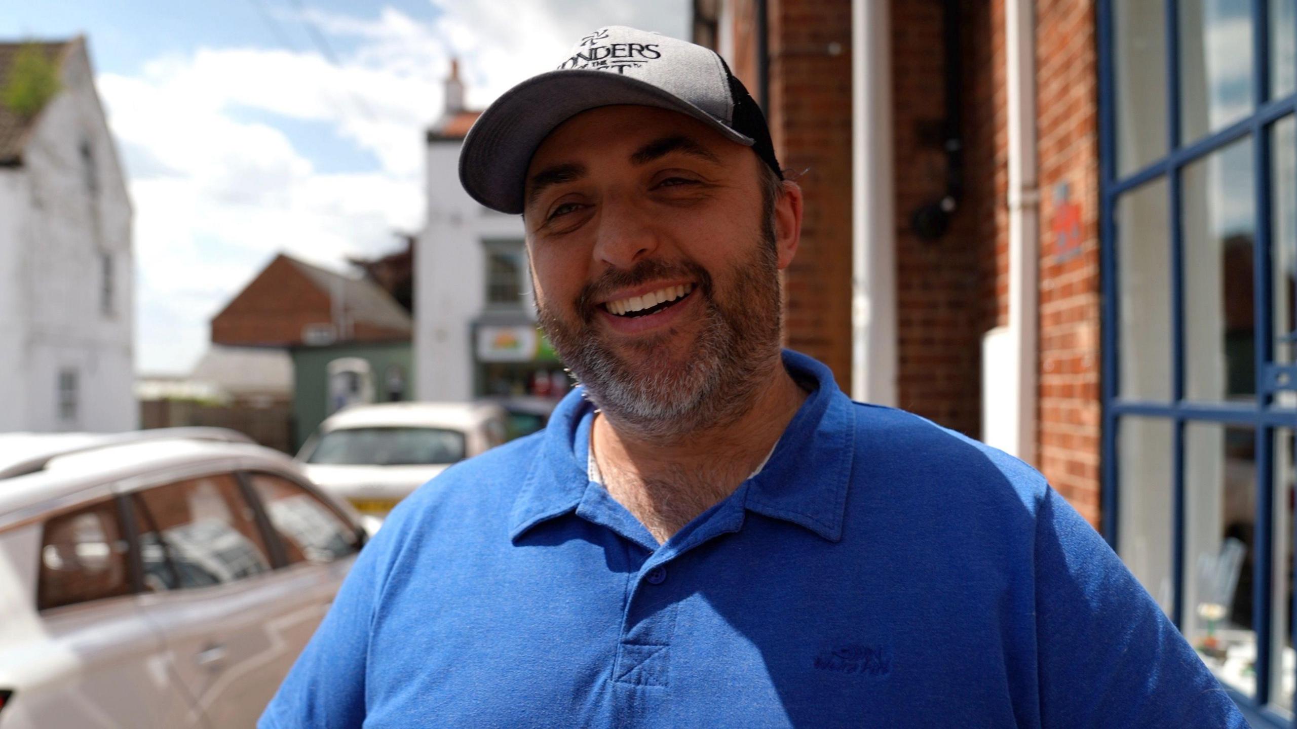 Brick Hut gaming centre owner Chris Allsopp is centre of the photo smiling at the camera. He is in his 40s and has tanned skin, dark eyebrows and a grey beard. He is wearing a grey baseball cap and blue polo shirt. To his left, there is a parked white car. To his right, there is a blue framed window and red brick wall of a building.