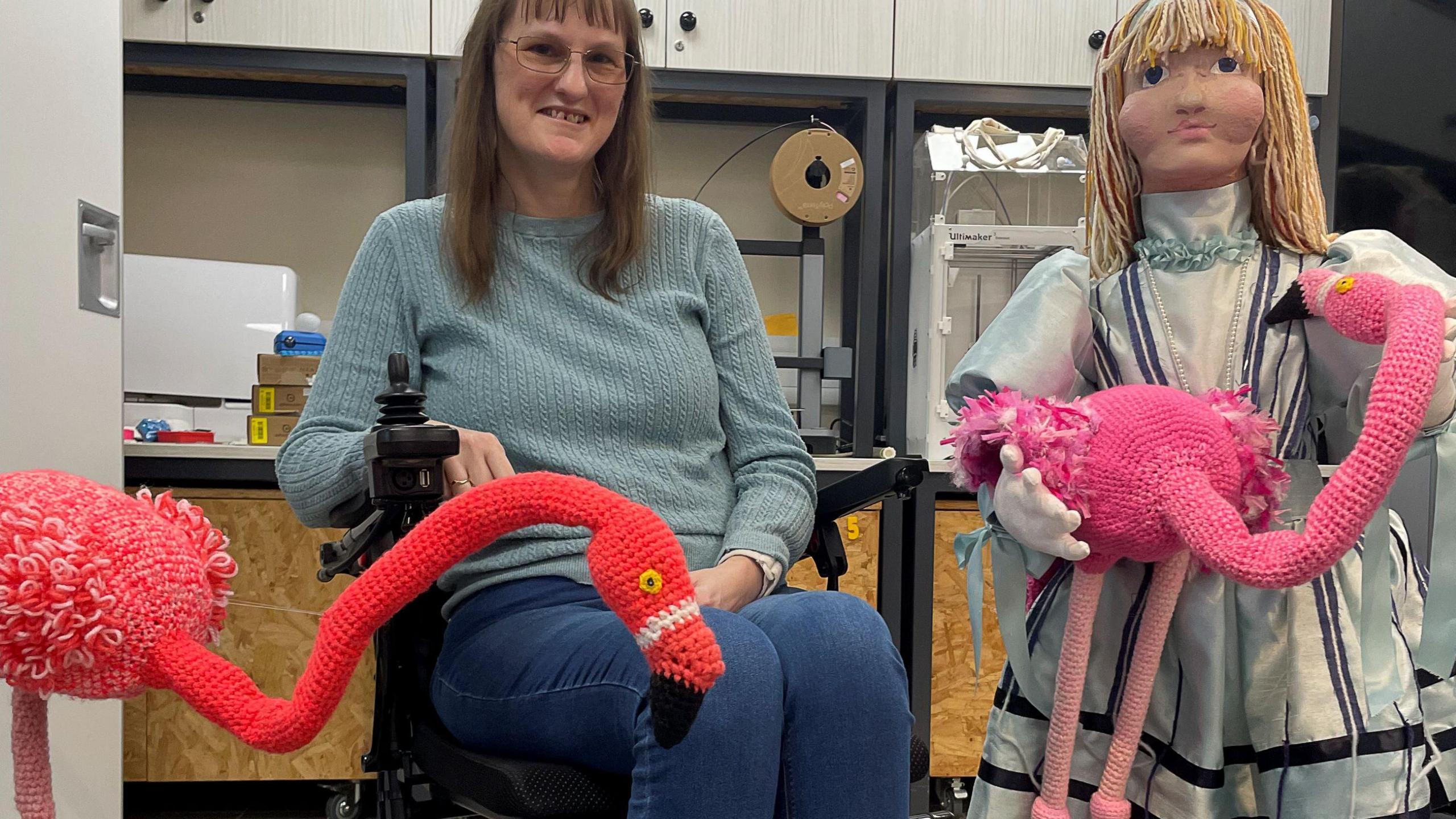 A woman wearing glasses, jeans and a jumper smiles at the camera. She is in a wheelchair and has a crafted orange flamingo next to her. Beside the woman is a knitted Alice who is holding a crafted flamingo.