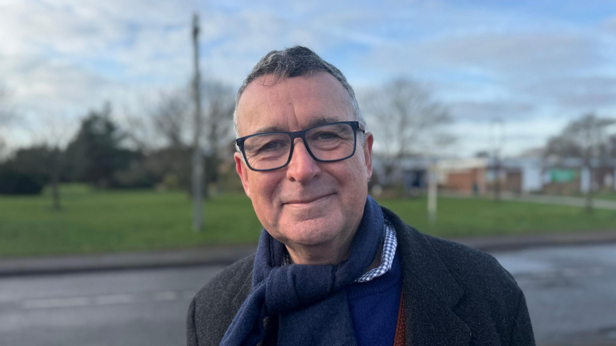 Sir Bernard Jenkin smiling straight at the camera. He is standing on a pavement opposite a community centre and is wearing a navy scarf, dark grey coat, and a royal blue jumper over a blue and white checked shirt.