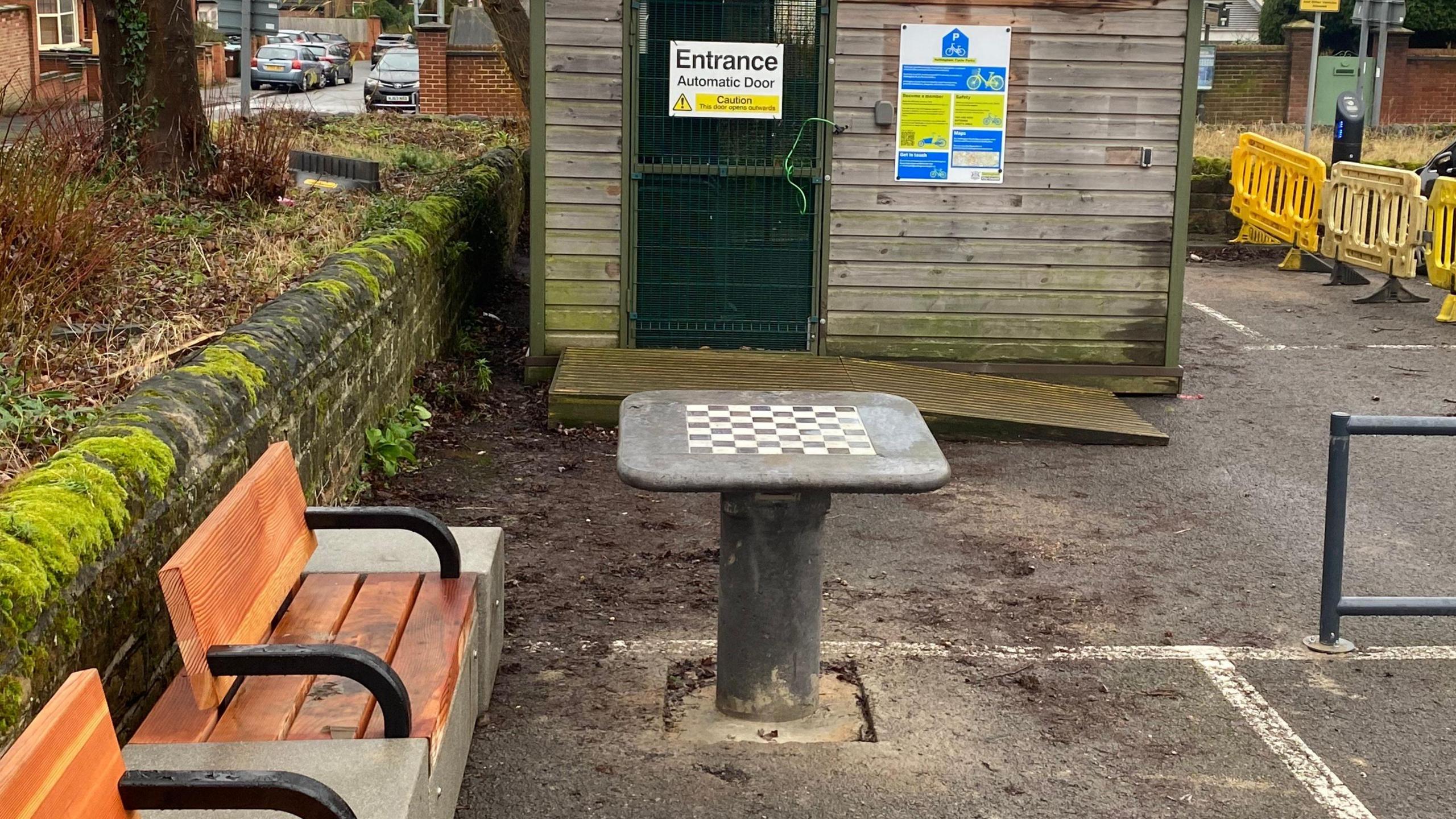 A grey table with a cheqeured design and bench in a car park 