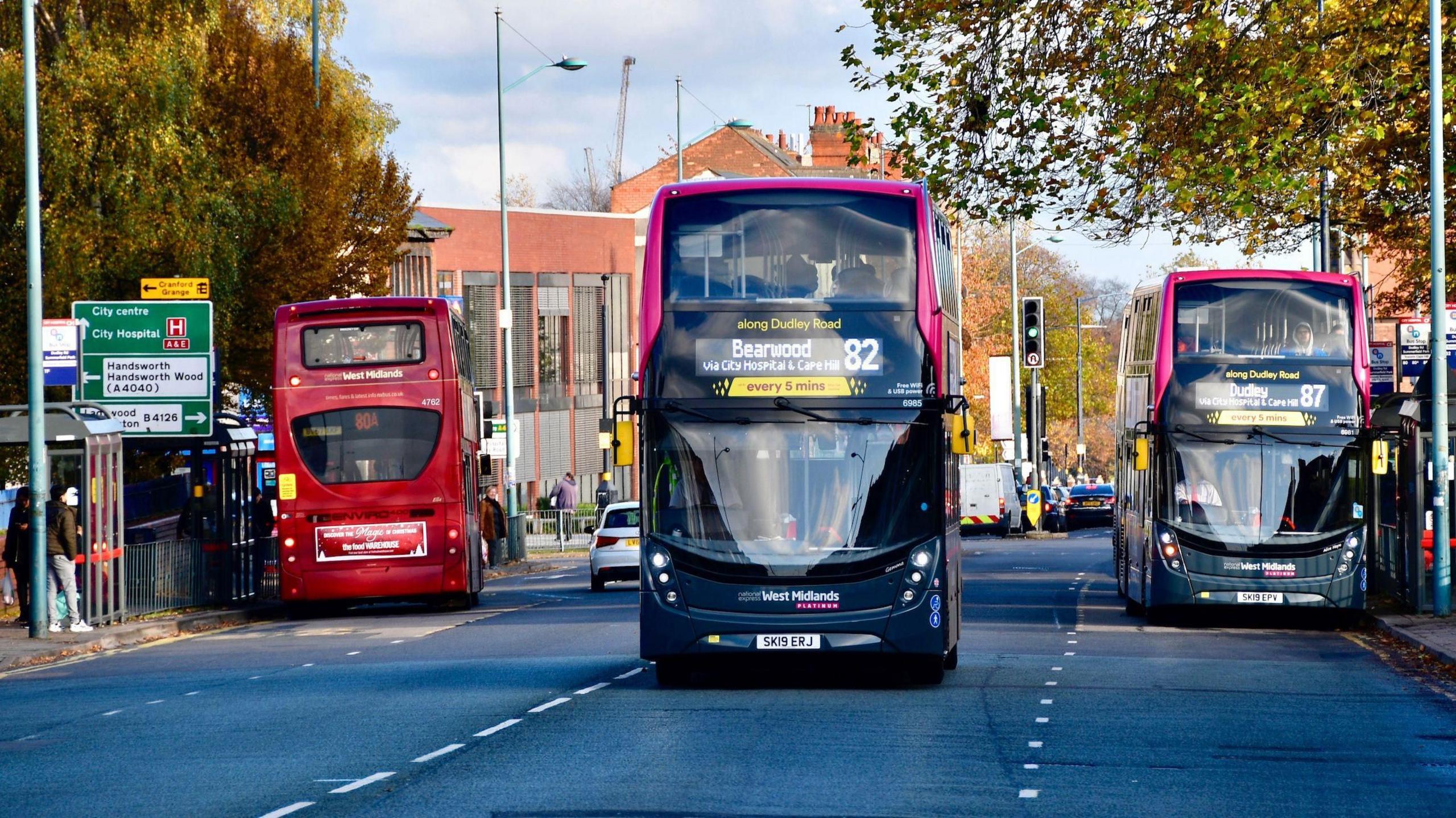 A three-lane road with three buses on it. A red bus is at a bus stop facing away from the camera. Another bus, with Dudley marked as its destination, is at a bus stop on the other side of the road facing the camera. Between them, a bus heading for Bearwood is driving towards the camera.