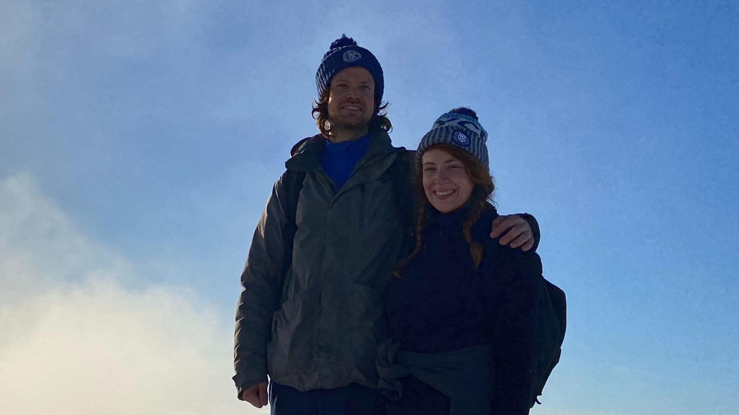Neil has his arm around Kat, who is wearing a woollen hat. They are standing against a blue sky