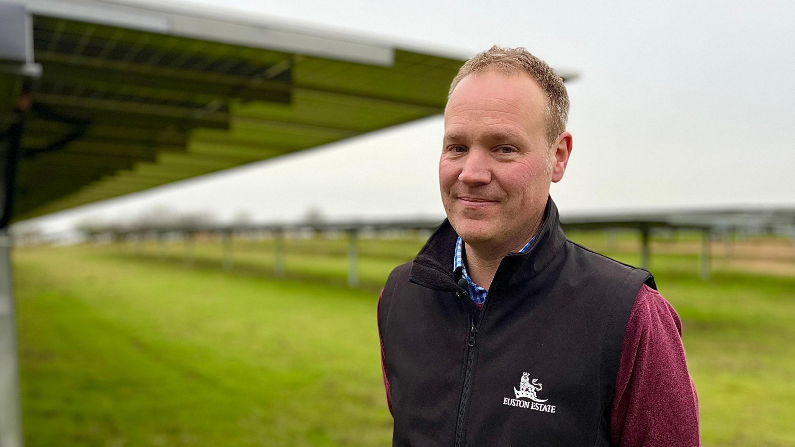 Robert Childerhouse looking down the camera while standing in front of rows of solar panels. He is smiling and wearing a black gillet with 'Euston Estate' and the estate's emblem - a crowned lion standing on a flag - embroidered on the chest. The solar panels appear to be mounted taller than Robert and have grass growing beneath them.  