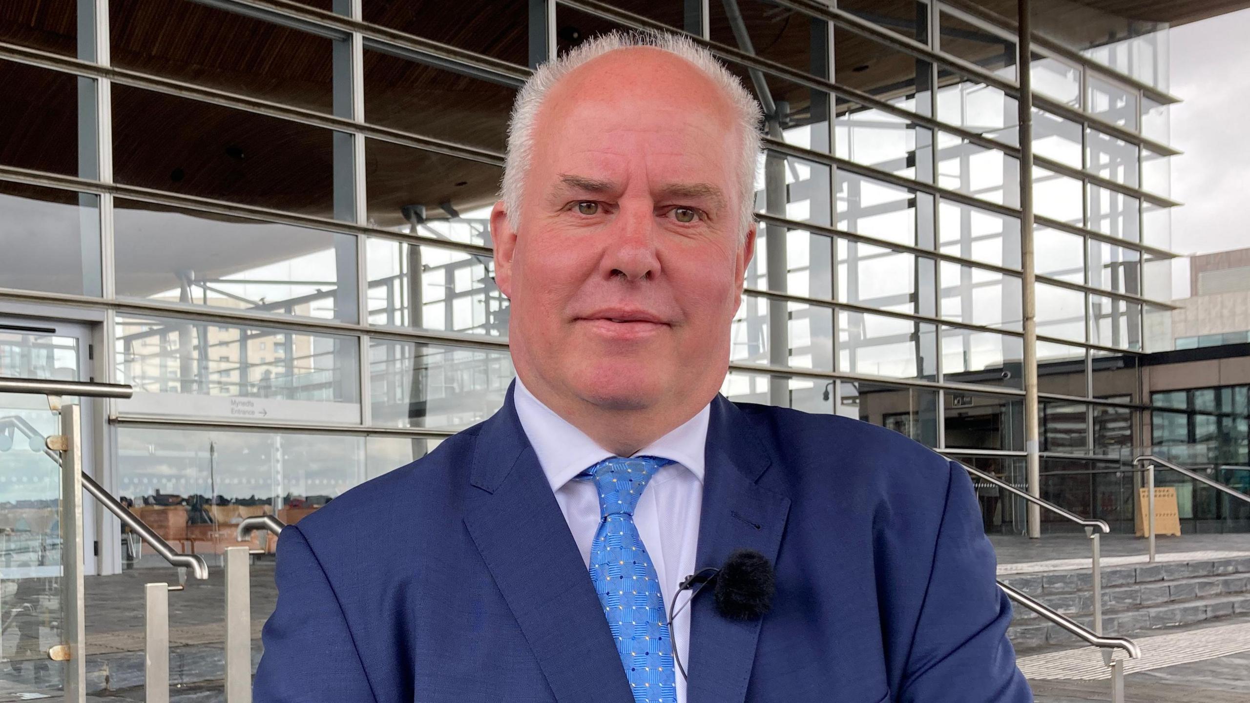 Andrew RT Davies wearing a dark blue suit, a light blue tie, a slightly purple shirt and a microphone stood on the steps in front of the Senedd building in Cardiff Bay.