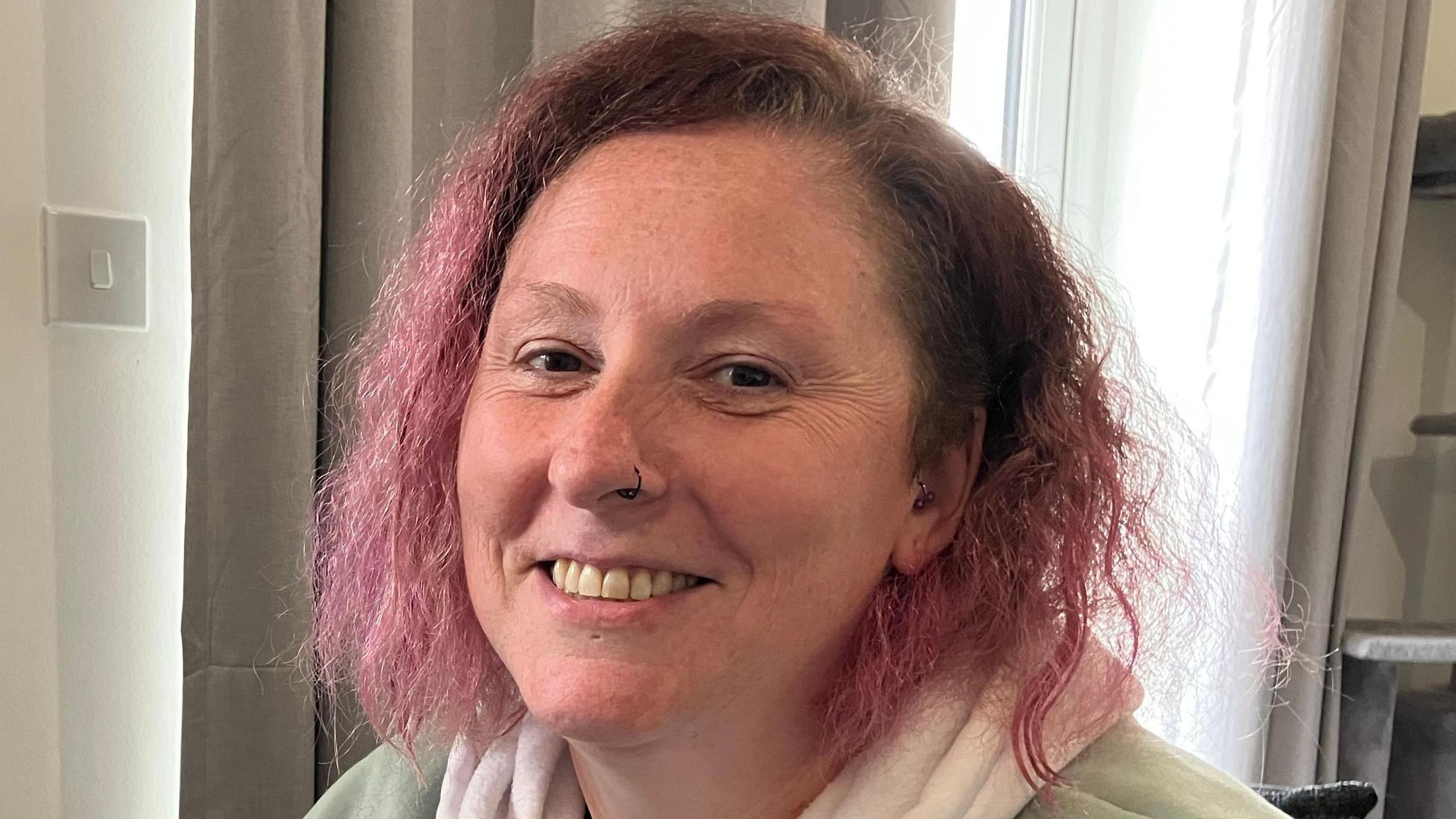 A woman with curly pink hair and a black nose ring smiles at the camera.