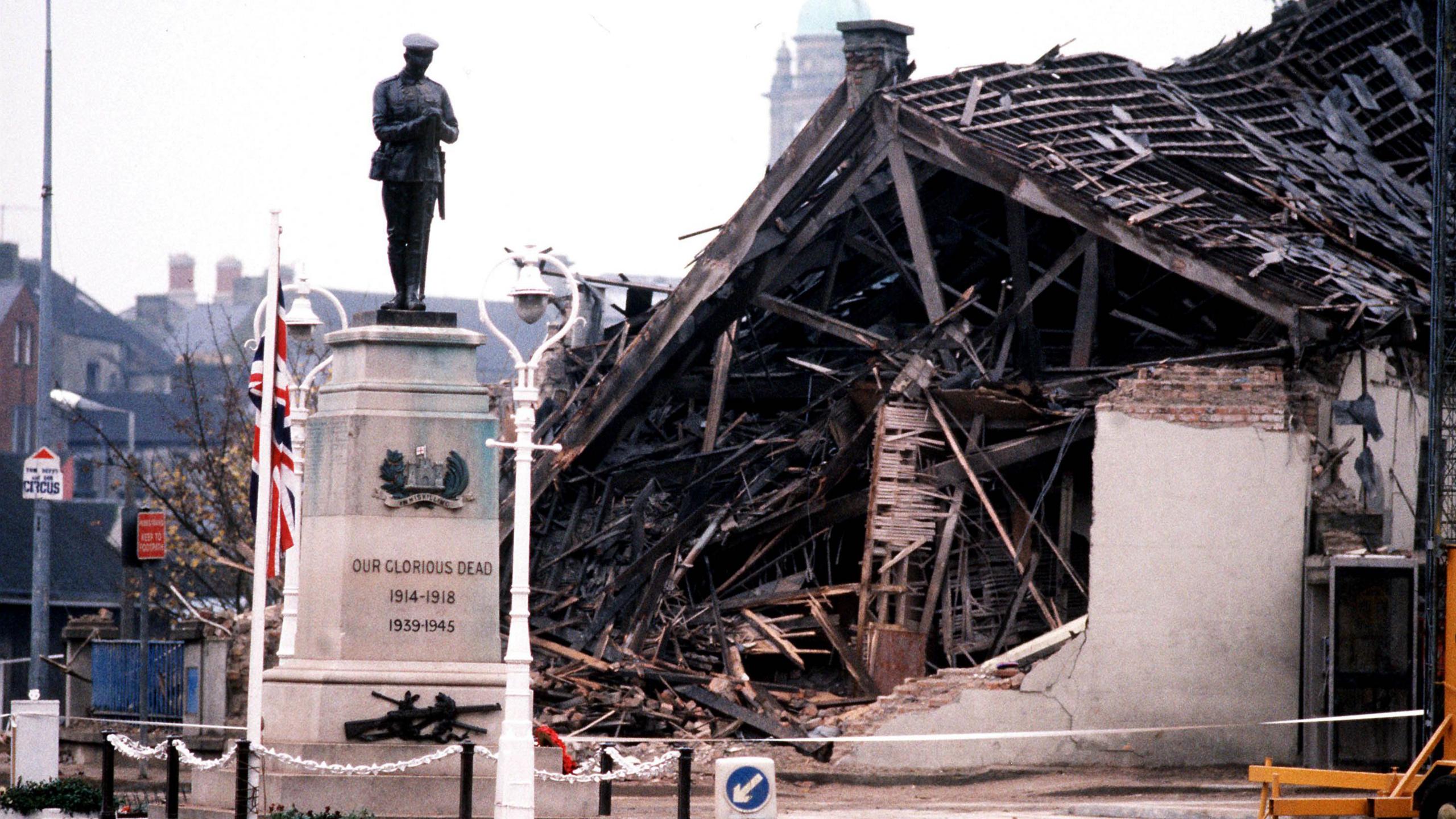 A photo of the cenotaph in Enniskillen, behind the statue is a building, wrecked due to an explosion by the IRA. The statue is of a single solider on top of a stone plinth, with a union jack beside it. 