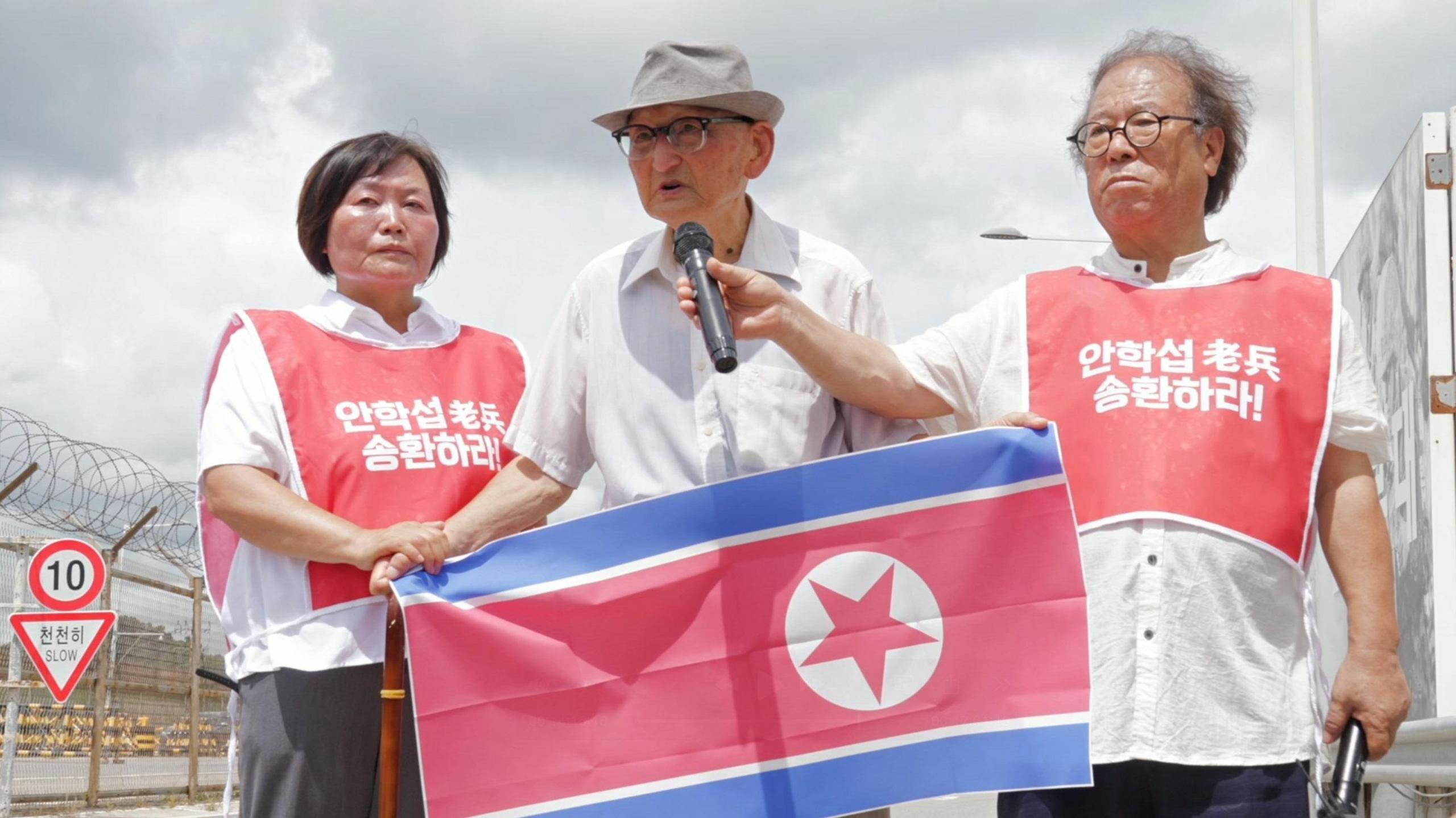 Ahn Hak-sop, wearing glasses, a hat, and a white shirt, holds a North Korean flag while two volunteers in red vests stand beside him - one holding a microphone for him, the other holding his right hand.