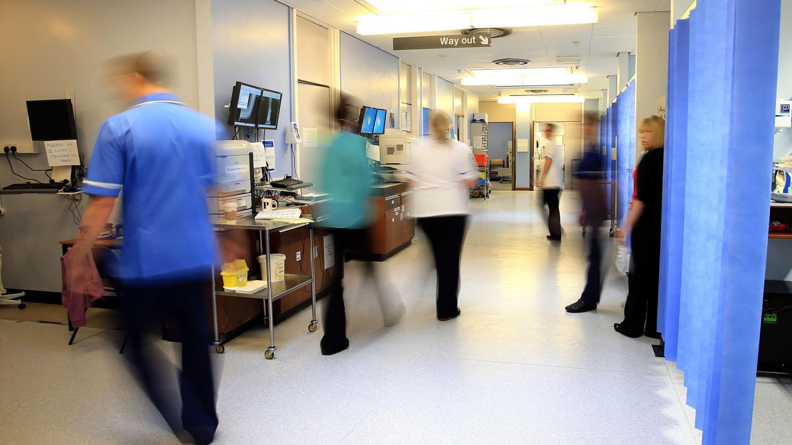 File photo of staff on a NHS hospital ward. All wearing scrubs of various colours, blue, white, green or black tops and all wearing black trousers. They are all blurred as pictured on the move in a ward with blue curtains and computers. 