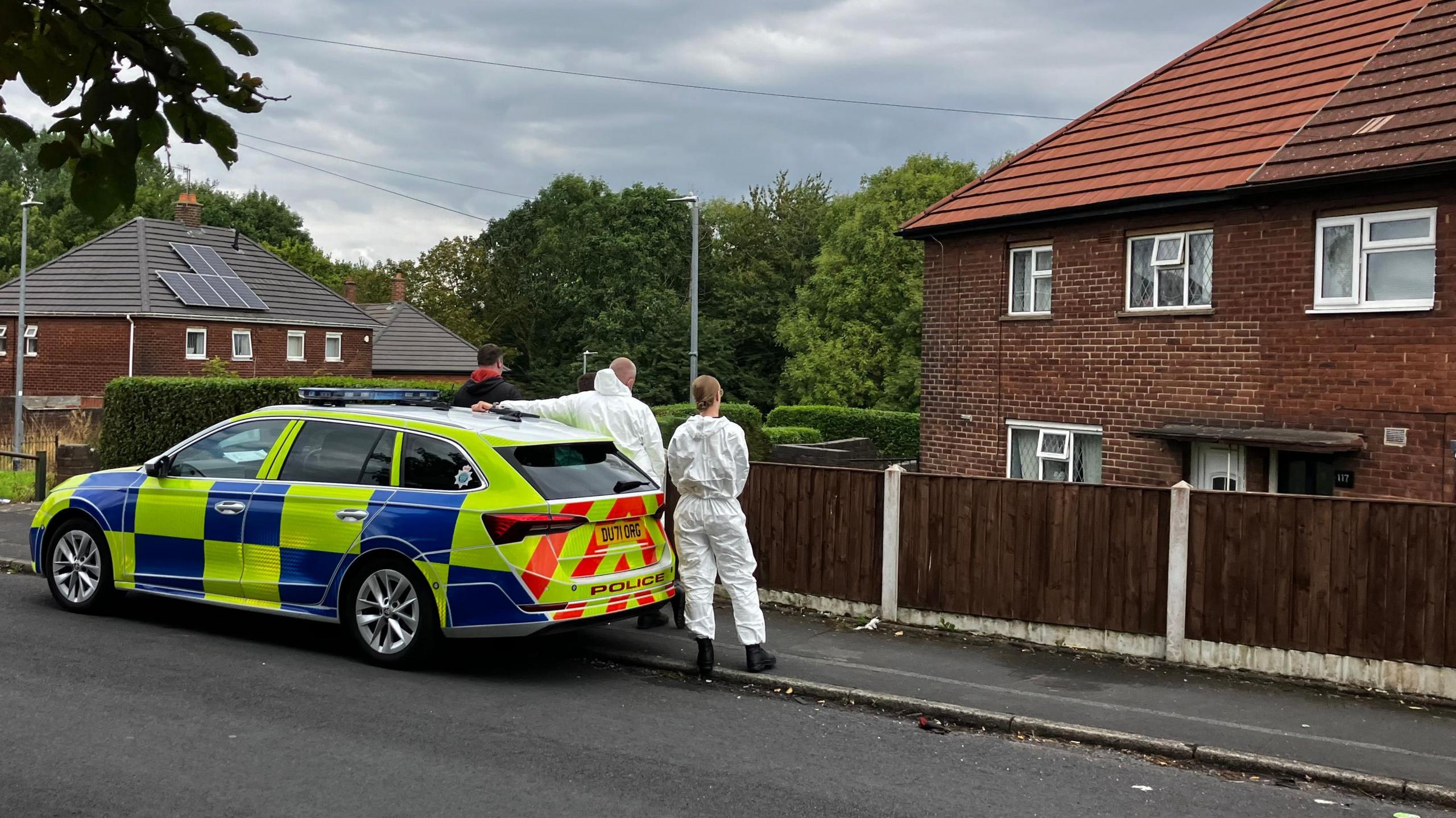 Two people in white overalls and another person in a black jacket with a red hood, stood next to a police car in front of a redbrick house with brown fence.