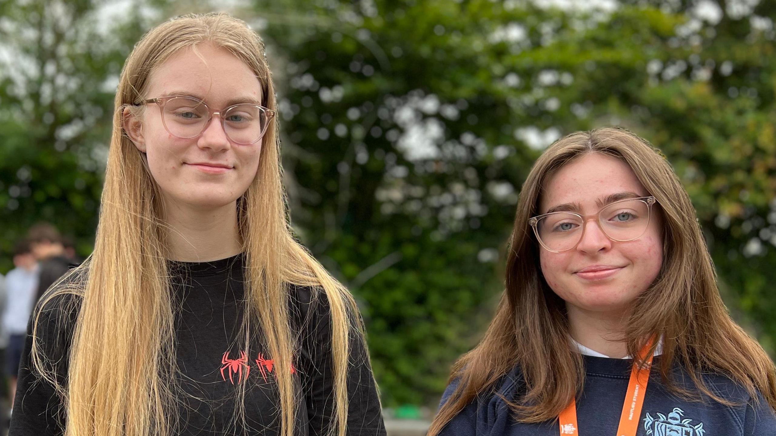 Chlodie and Lucy are standing outdoors on a paved area with trees in the background. Lucy is on right and has long, straight hair and is wearing a black T-shirt. Chlodie is on the right and has shoulder-length hair and is wearing a dark blue sweatshirt with an orange lanyard around their neck, which holds an ID card. In the background, several other people are gathered near a go-kart.