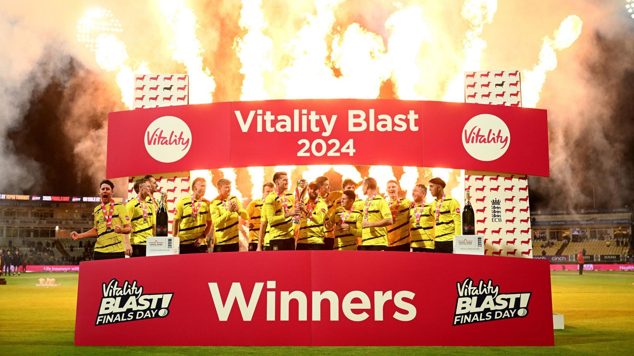 Gloucestershire cricketers celebrate winning the T20 blast final against Somerset at Edgbaston. They are standing on a plinth with fireworks going off overhead, and a large sign with red background and white writing says "T20 blast champions"