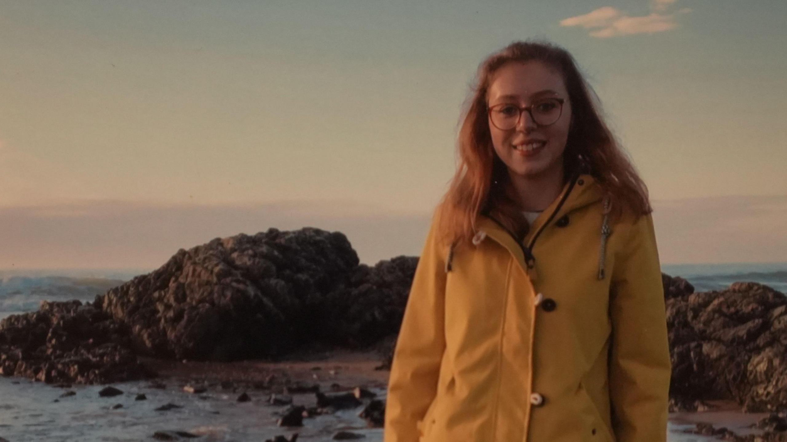 Mared Foulkes looking at camera standing on the Anglesey coast wearing a yellow coat