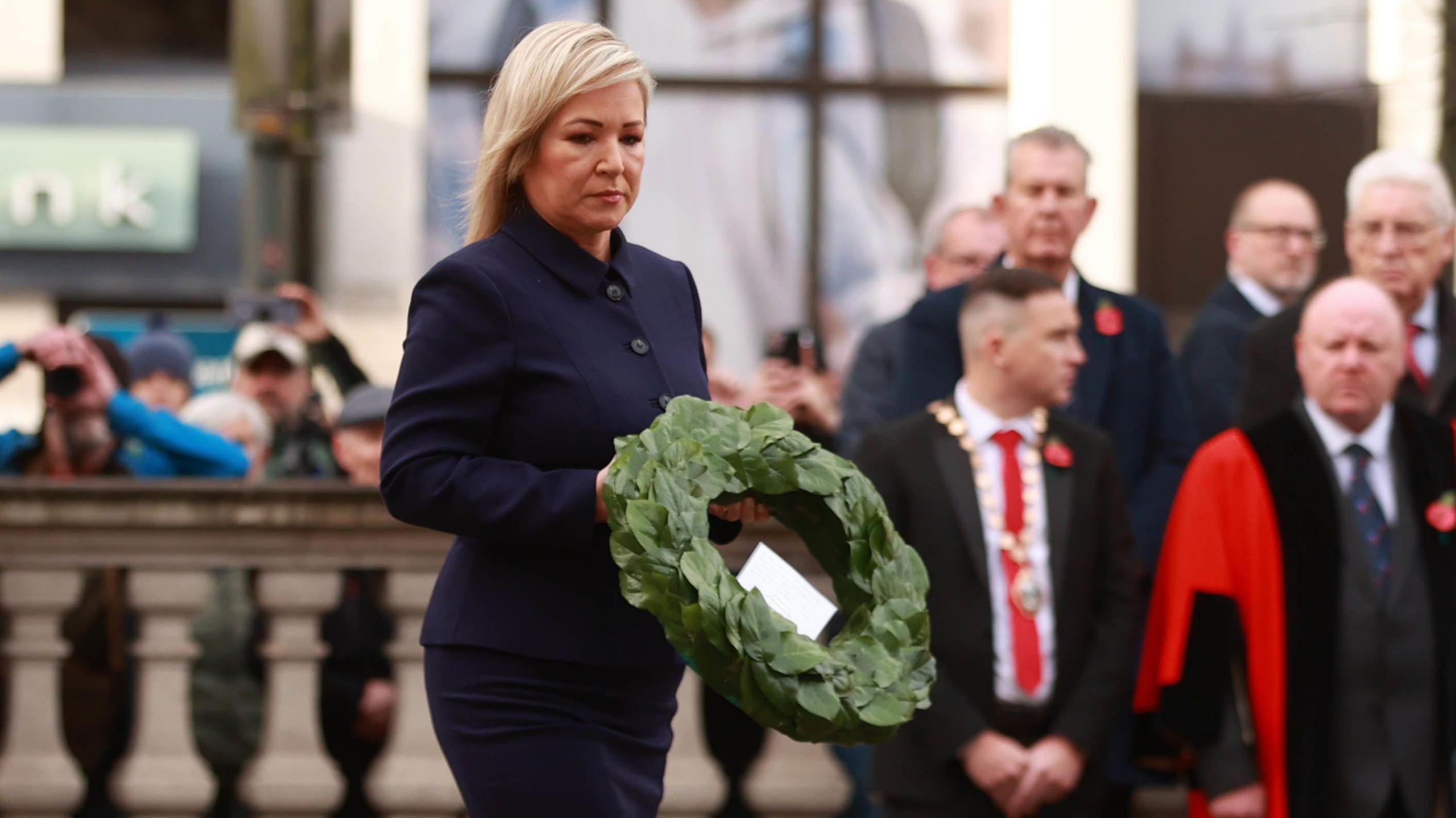 Michell O'Neill wearing a blue jacket carrying a green wreath. Behind her some men stand out of focus wearing suits and poppies. To her left behind stone columns some people hold up cameras and phone to take photos.