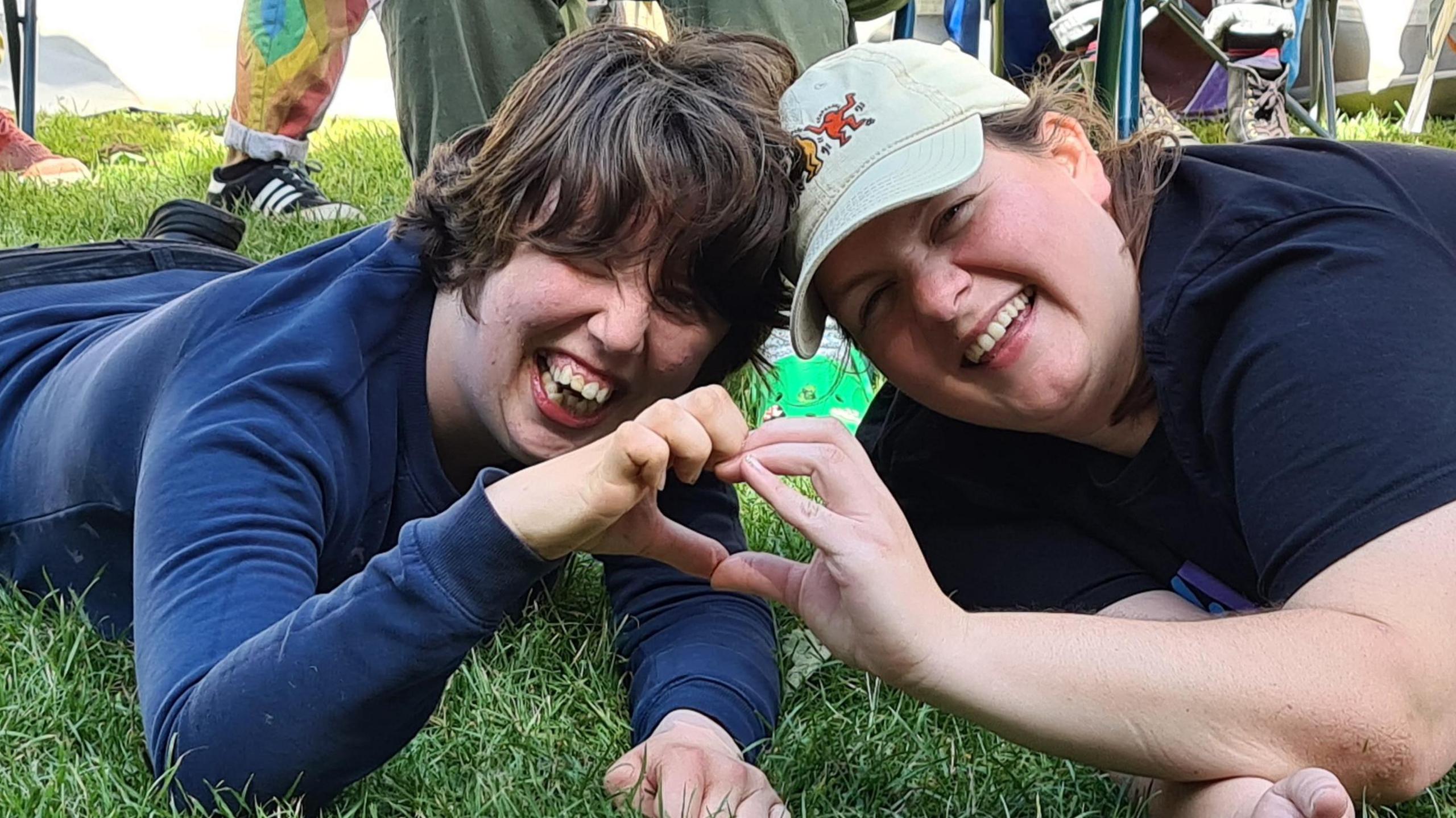 Two young people smile broadly at the camera as they lie on some grass and make a heart with their hands.