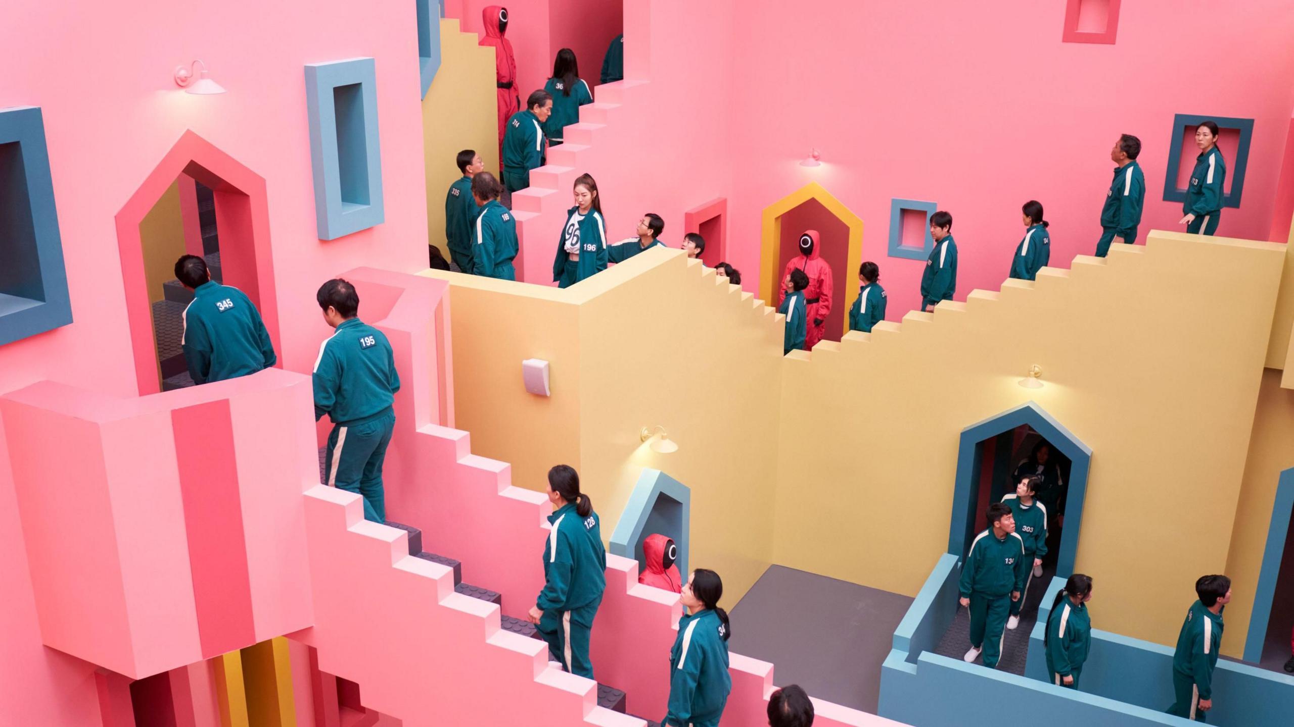 Contestants in dark blue track suits walk up and down the distinctive staircase which is painted pink, blue and yellow.
