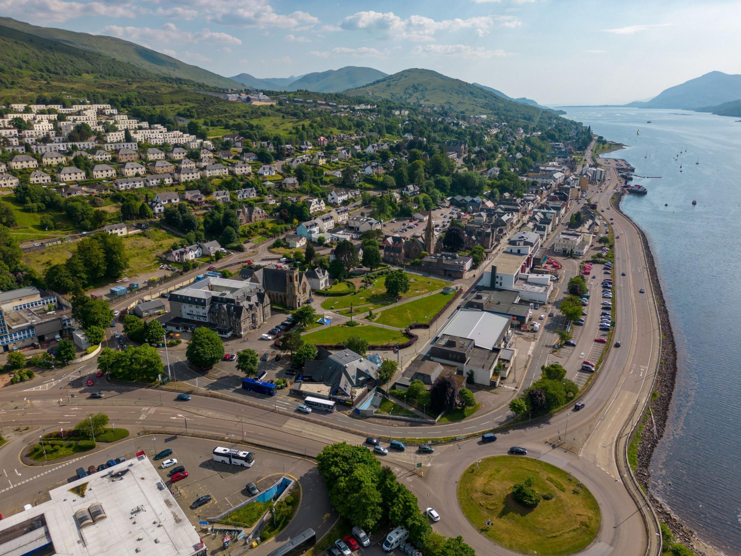 An aerial view of Fort William in Lochaber. Rows of houses and businesses are on a hillside among trees and also line a shore of Loch Linnhe.