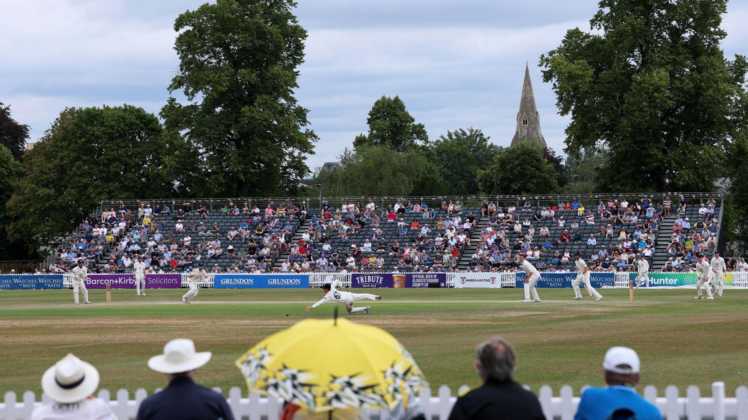 Crowds watch on at The College Ground in Cheltenham as Gloucestershire cricketers play Middlesex at the annual festival in the town. In the background is a temporary grandstand with green seats, and beyond that trees and a church spire are visible