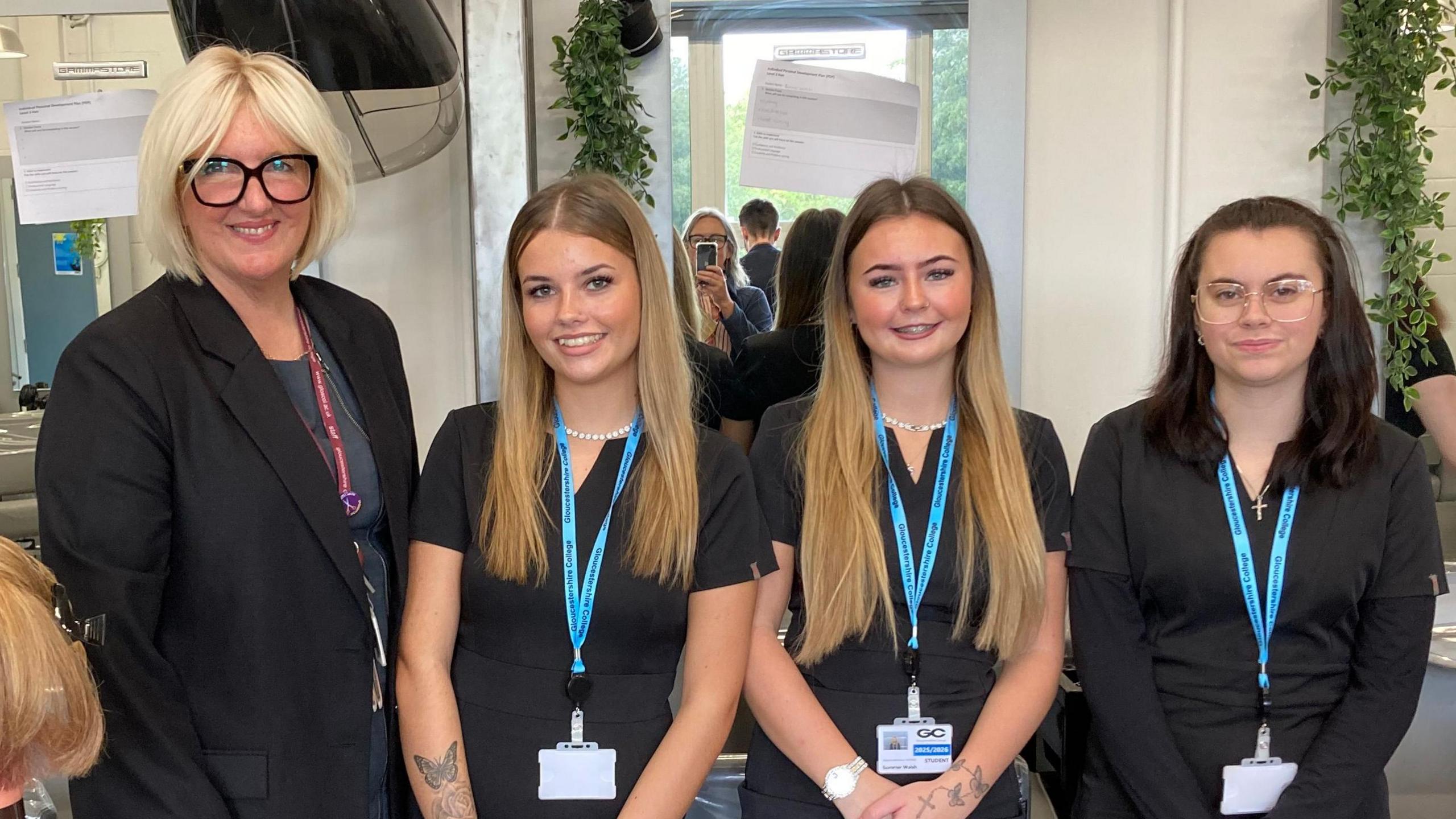 Three young girls standing next to their female college tutor. The tutor has short blonde hair and is wearing glasses with thick black frames. The two girls to her left have long blonde hair. The girl on the end of the line has long dark hair. All of them are wearing black and have lanyards around their necks.
