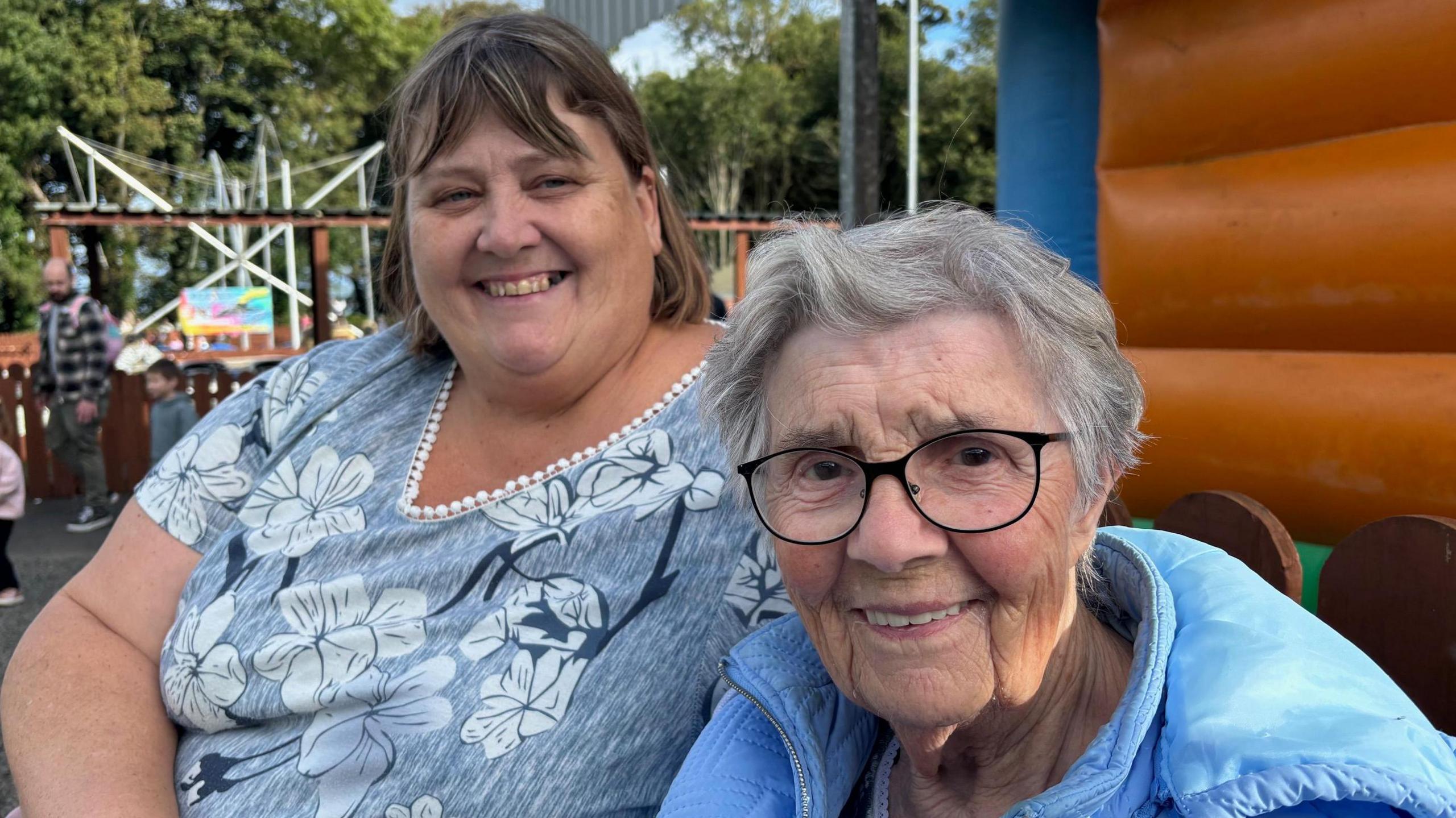 Two ladies smiling at camera while at the fun zone at Carnfunnock Country Park . One is wearing a blue patterned top, the other has short grey hair and glasses and is wearing a sky blue hooded and padded coat.