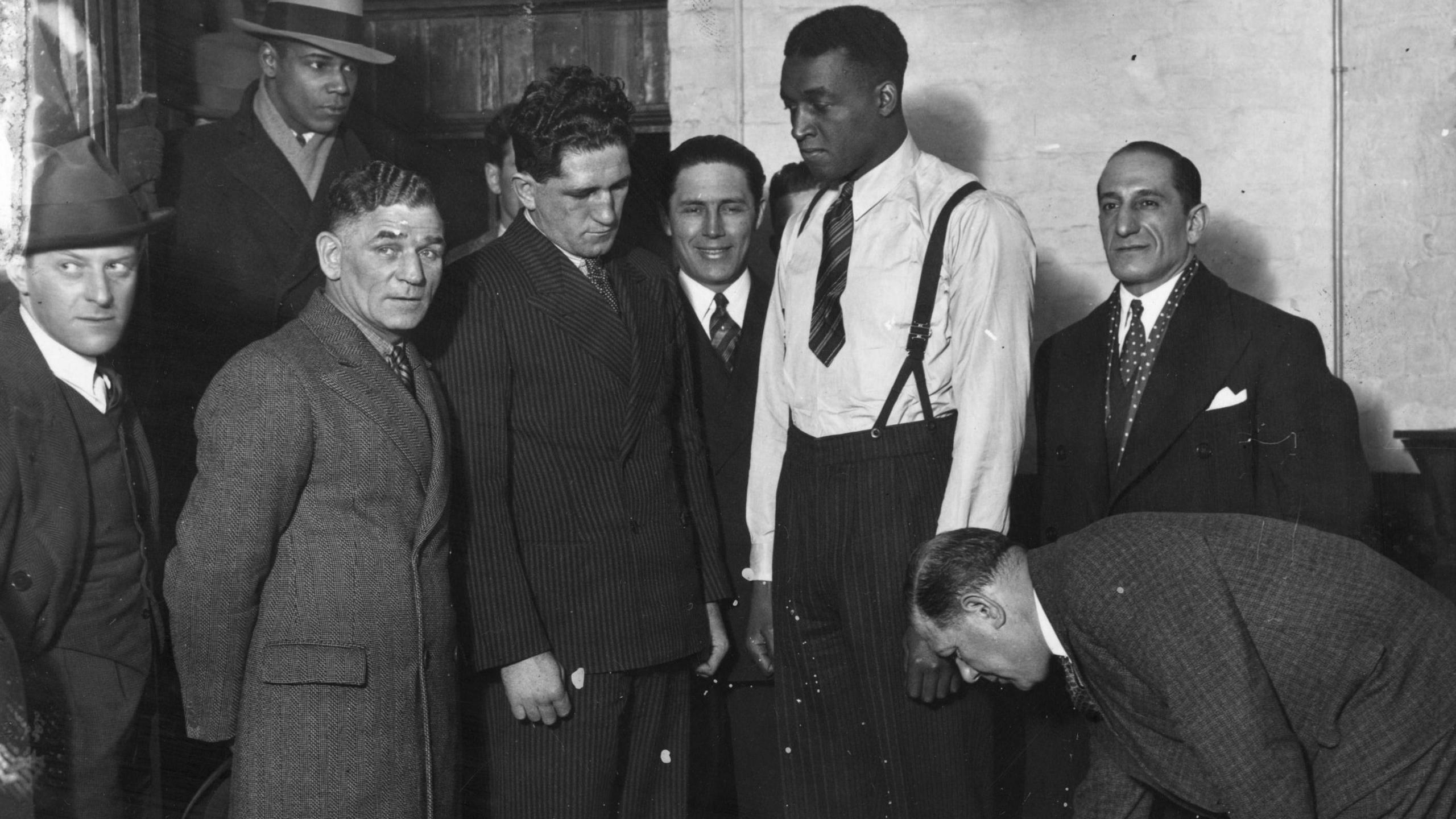 Len Johnson, wearing a white shirt and striped tie, stands on scales and is surrounded by men wearing suits and overcoats