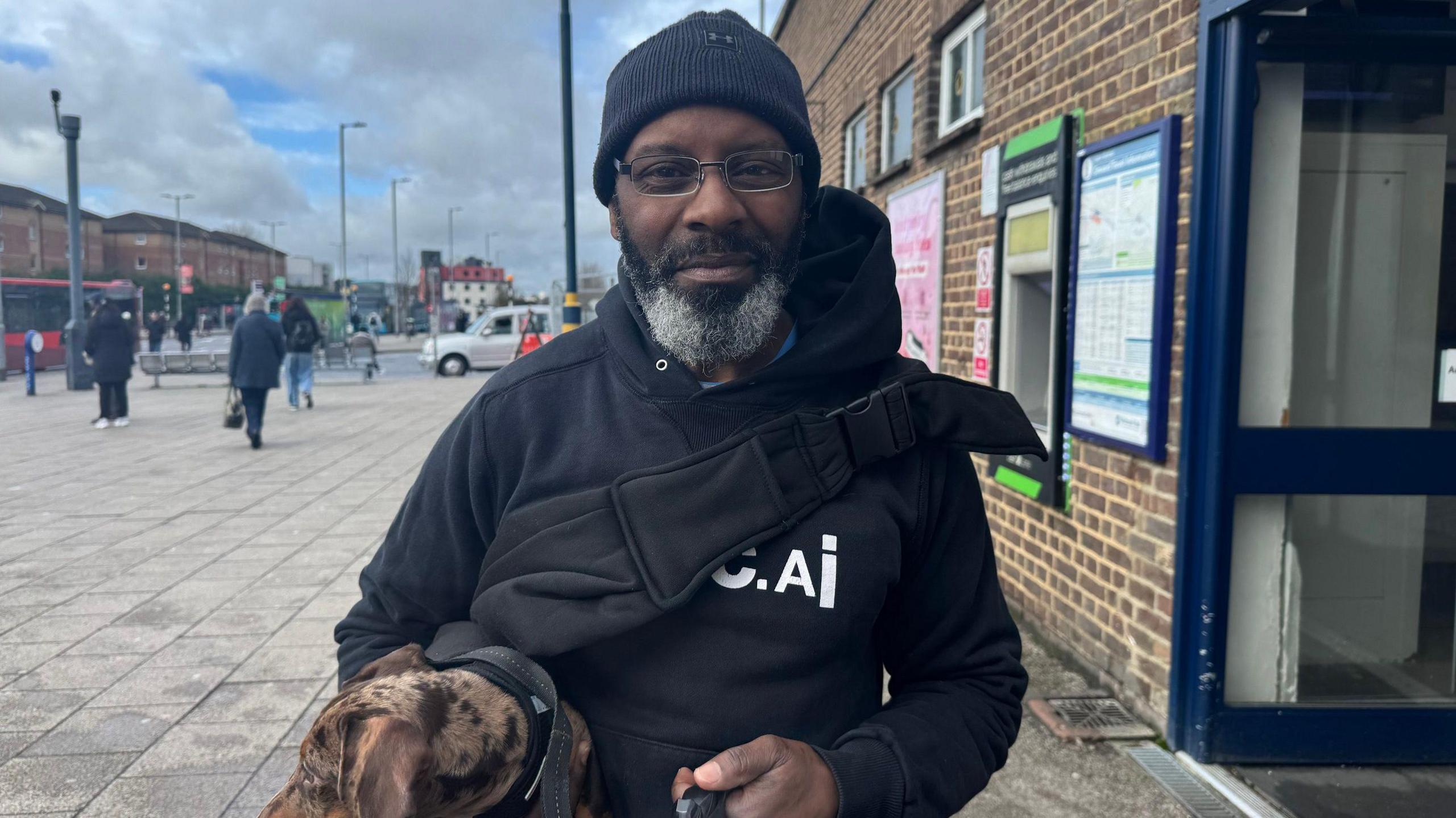 Howard Malcolm pictured outside Luton train station, with lamp posts and people behind him. His is holding a brown speckled dog and wears a black beanie hat, glasses, a black hooded jacket and the black strap of a bag across his chest.