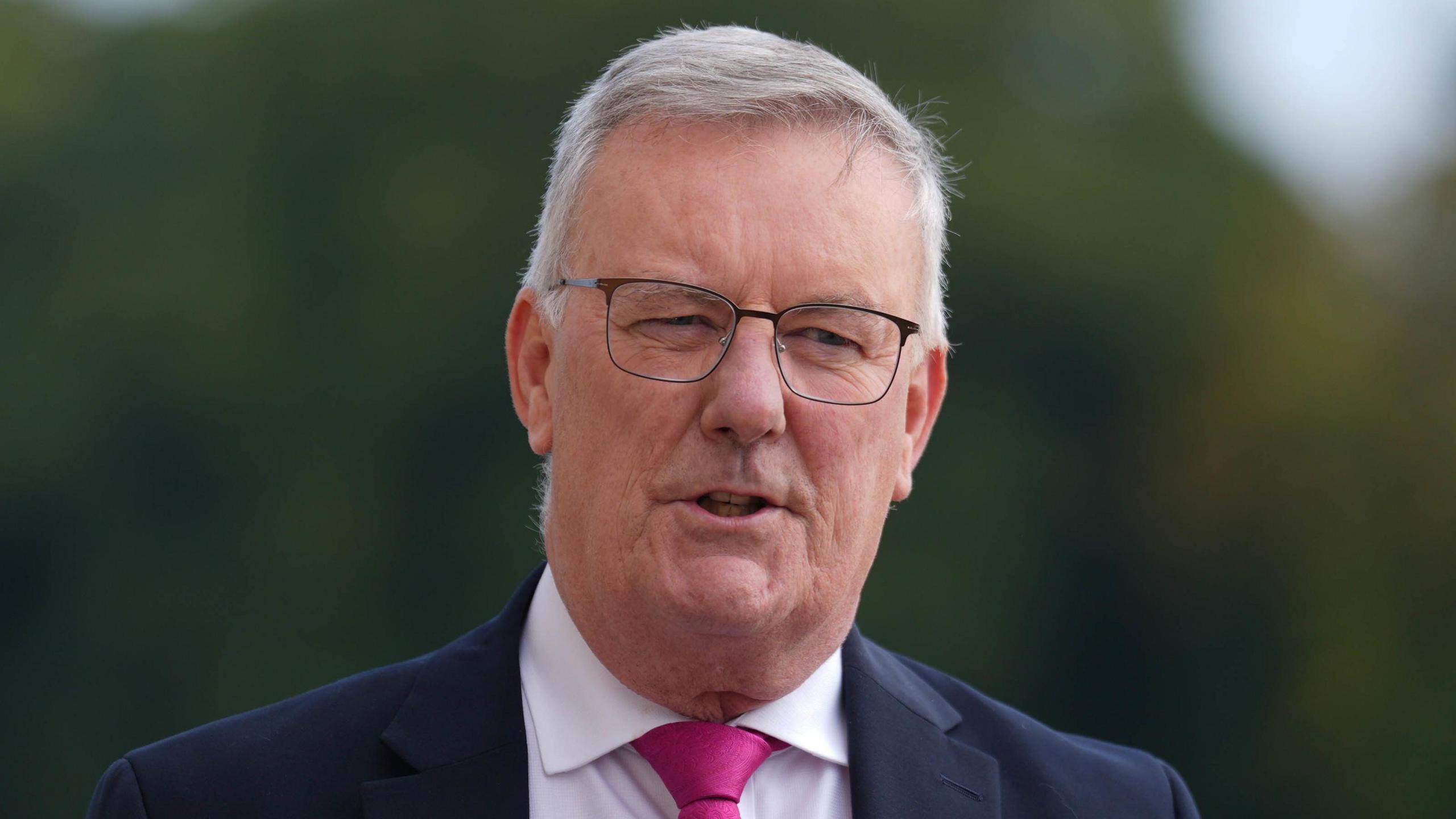 Mike Nesbitt, who has short grey hair, is standing outside. There are green trees behind him. He is wearing a navy suit, white shirt and pink tie. He is wearing glasses.