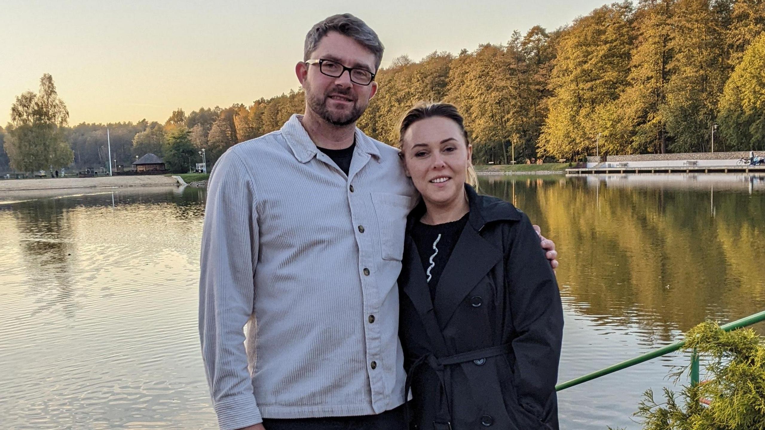 Robbie Anderson stands with his wife in front of a lake. He wears a grey, cord, button-down jacket and glasses and has his arm around his wife who wears a dark grey belted trench coat.