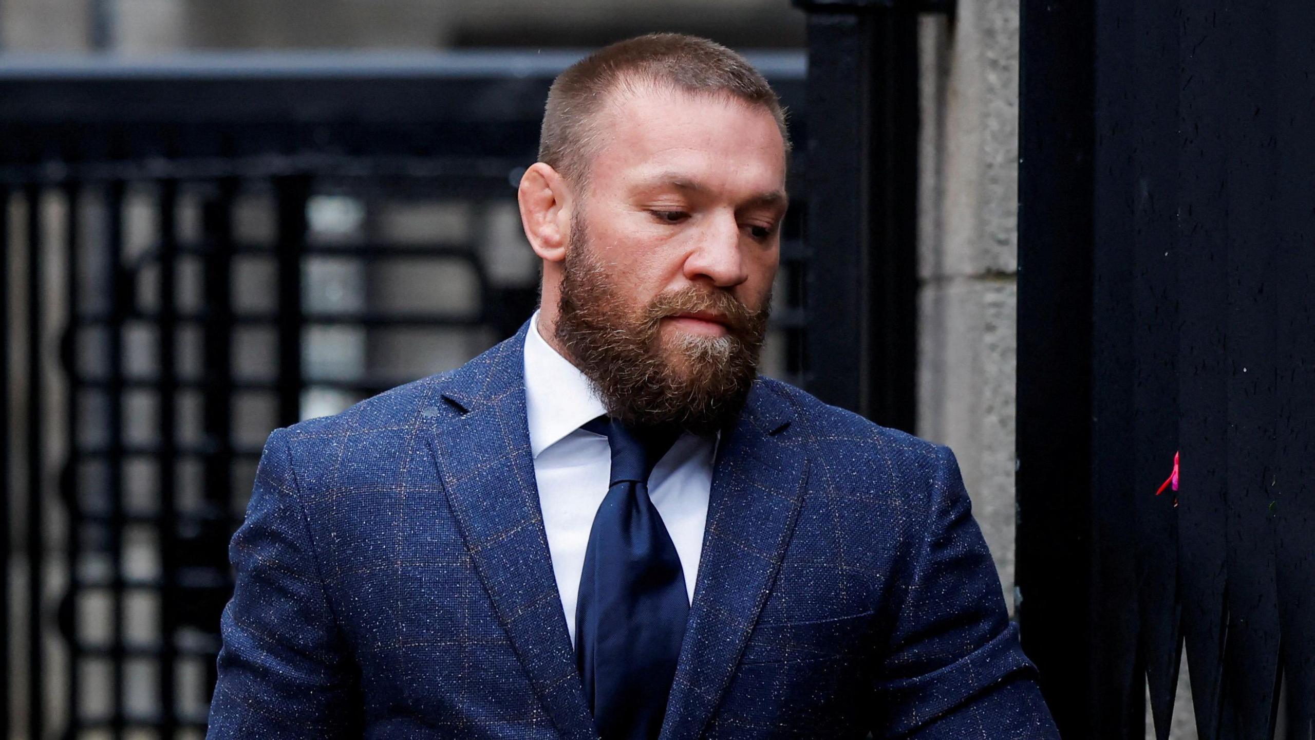 Conor McGregor, pictured from the chest up, wearing a navy tweed suit, a white shirt and a navy tie. He has tight brown hair and a brown beard. He is looking towards the ground. 