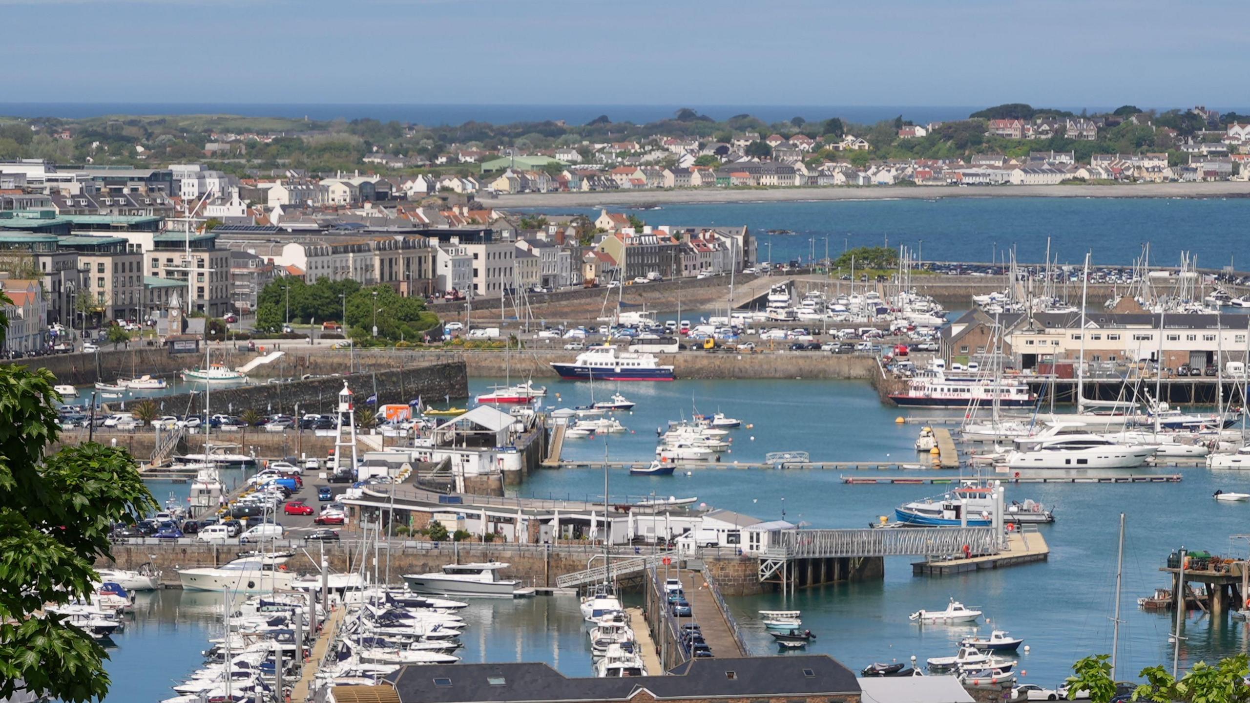 A photo showing the view across St Peter Port harbour to houses on the shore beyond, marina berths with boats moored at them are visible in the foreground and in the middle of the image which has been taken on a sunnny day