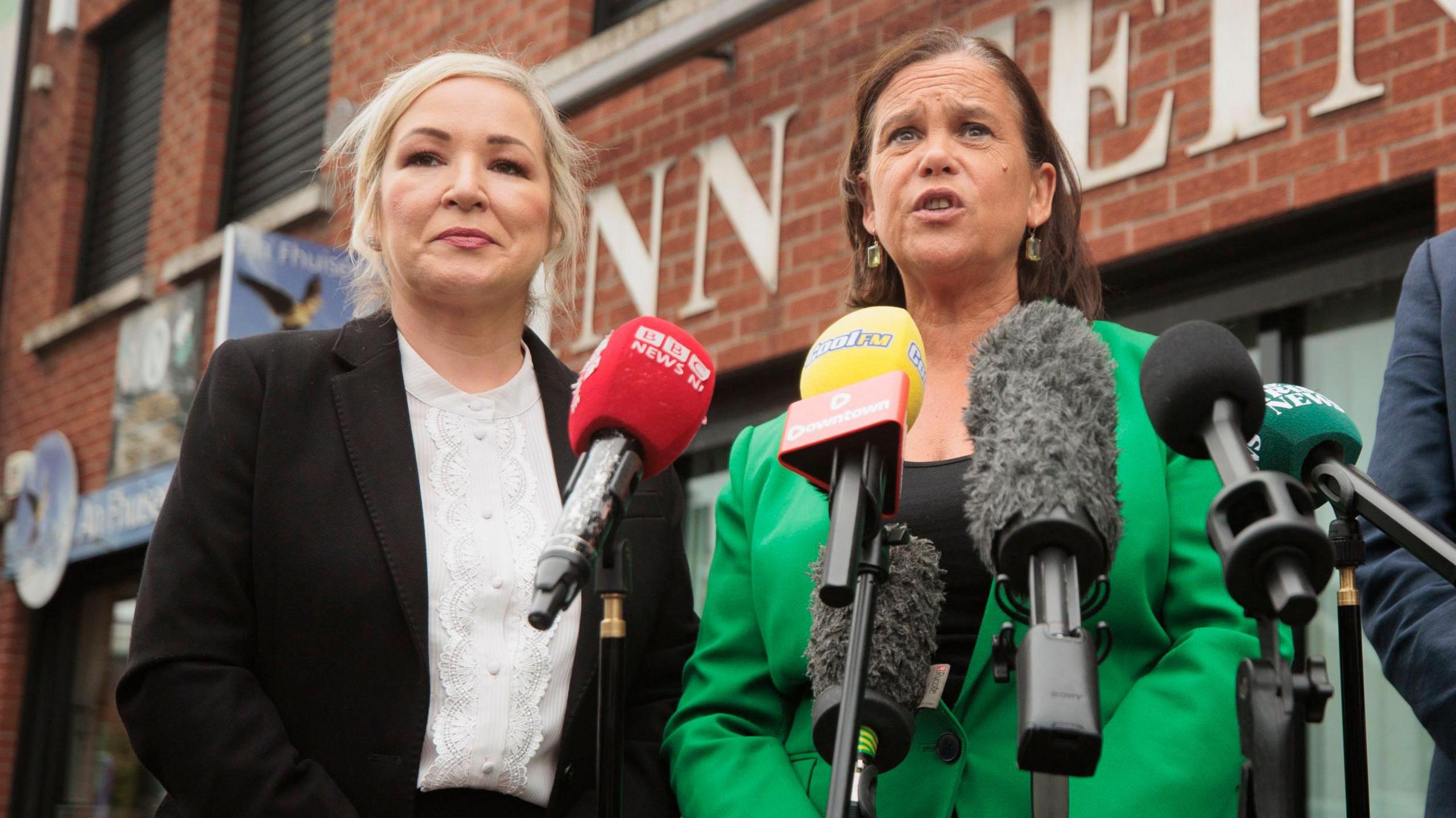 Michelle O'Neill, a woman with shoulder-length blonde hair, standing beside Mary Lou McDonald during a Sinn Féin press conference.  McDonalds has short, dark hair andis wearing a black top and a bright green blazer.  O'Neill is weaing a white blouse and black suit jacket. 