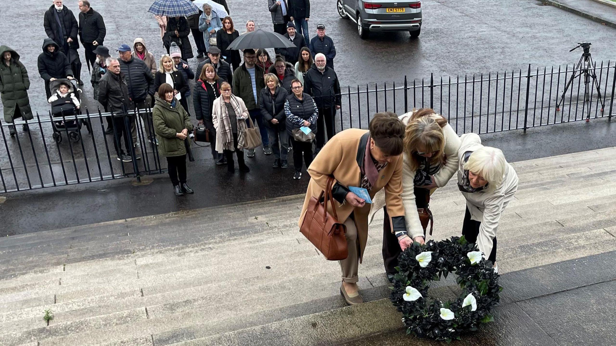 3 ladies lay a wreath on the steps of Stormont, the women on the left has brown hair, wearing a brown coat and nrown leather bag. The lady in the middle has brown hair, wearing a white coat and glasses. The lady on the right has white hair, wearing a white coat.