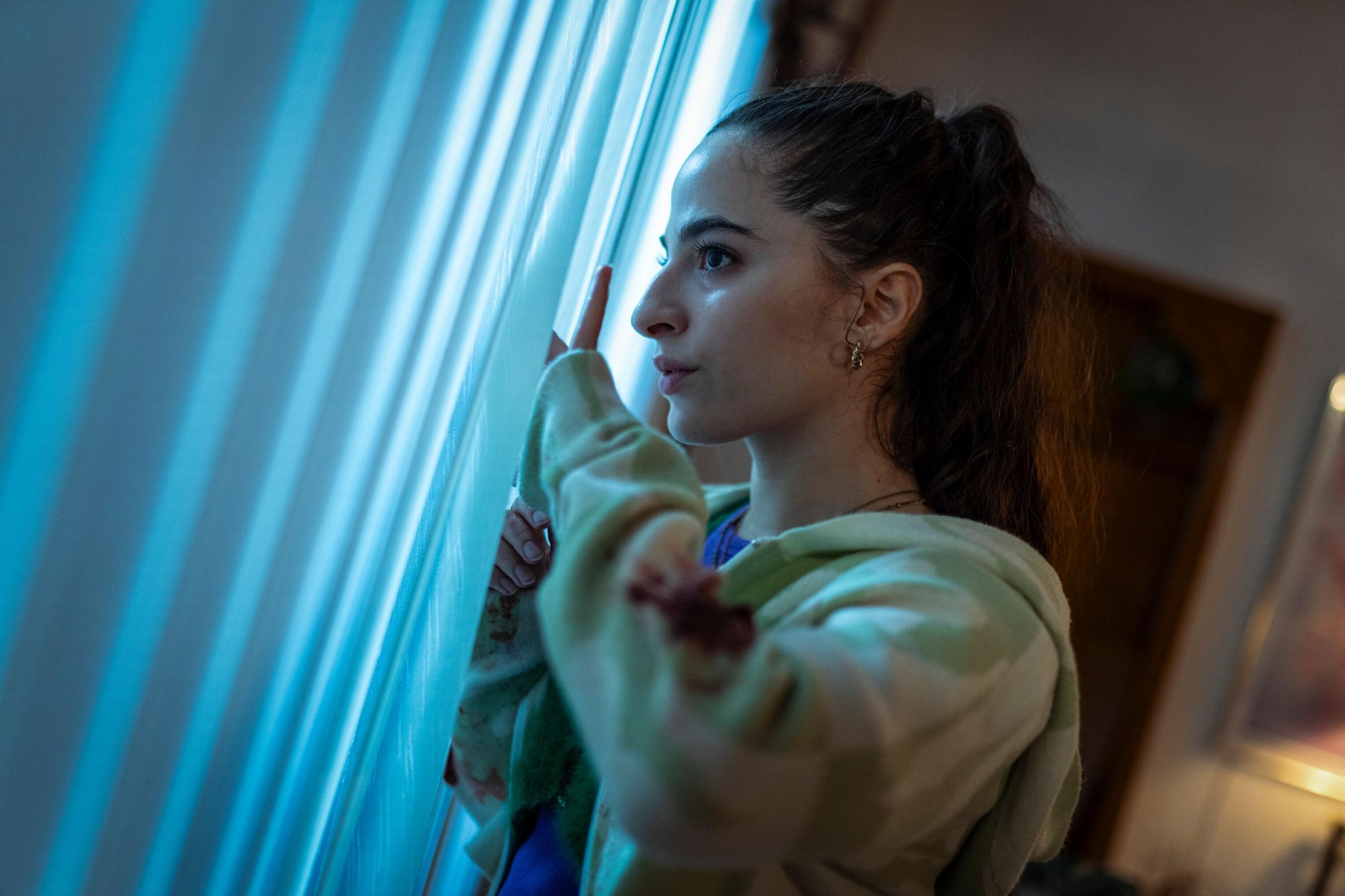 A young woman looks nervously out of a window through blinds.