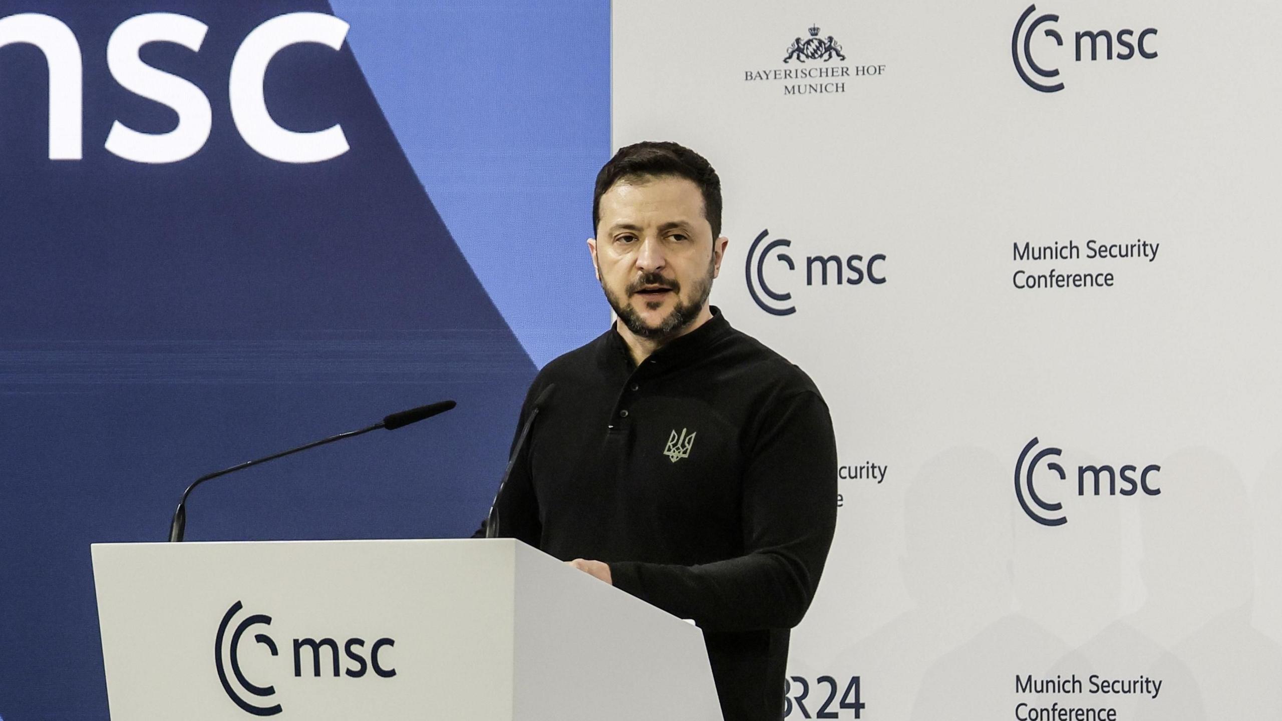 Volodymyr Zelensky stands at a lectern at the Munich Security Conference as he gives a speech. He is wearing a black fleece with the symbol of the Ukrainian Defence Forces embroidered on the breast.