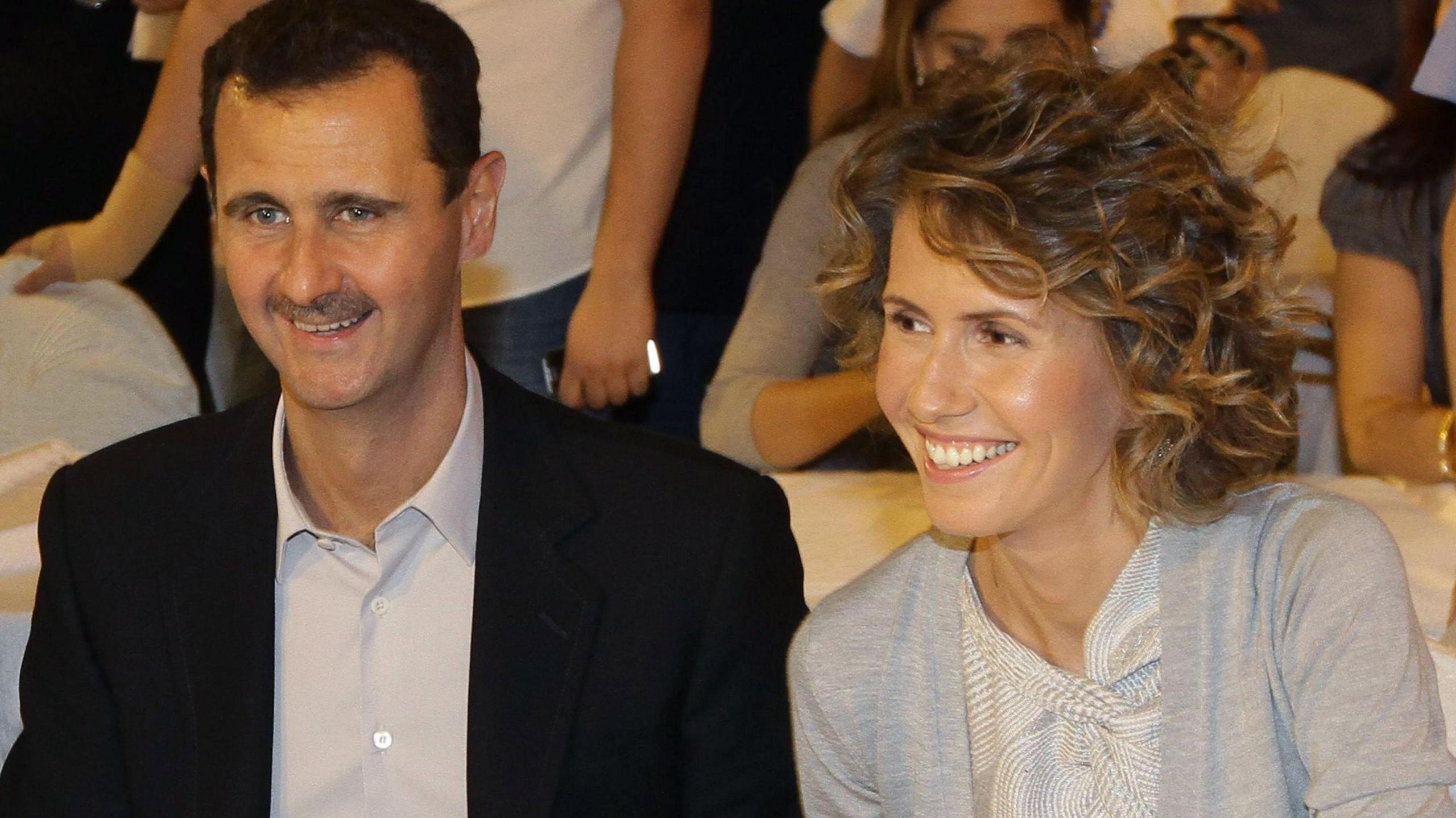 A smiling Bashar al-Assad in an black suit with no tie, sits next to a smiling Asma al-Assad in a cardigan 