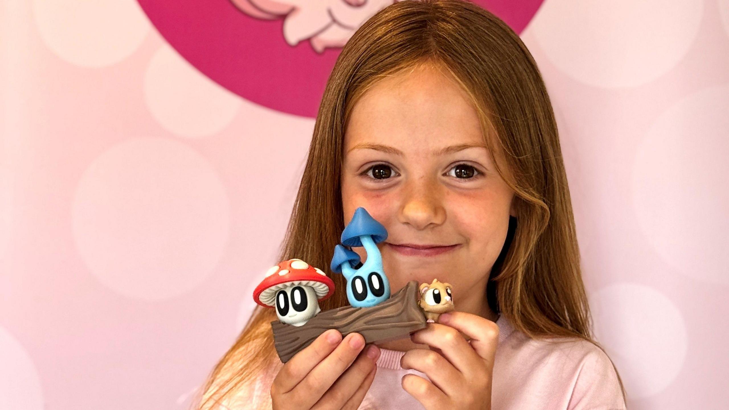 A girl with long brown hair, parted in the middle, smiles at the camera while holding a plastic toy in her hands, level with her face. The toy is a log, with a squirrel or mouse-like character poking is head out of one end. On the top of the log are two toadstools, one red and white, one blue. All characters have large cartoonish eyes. 