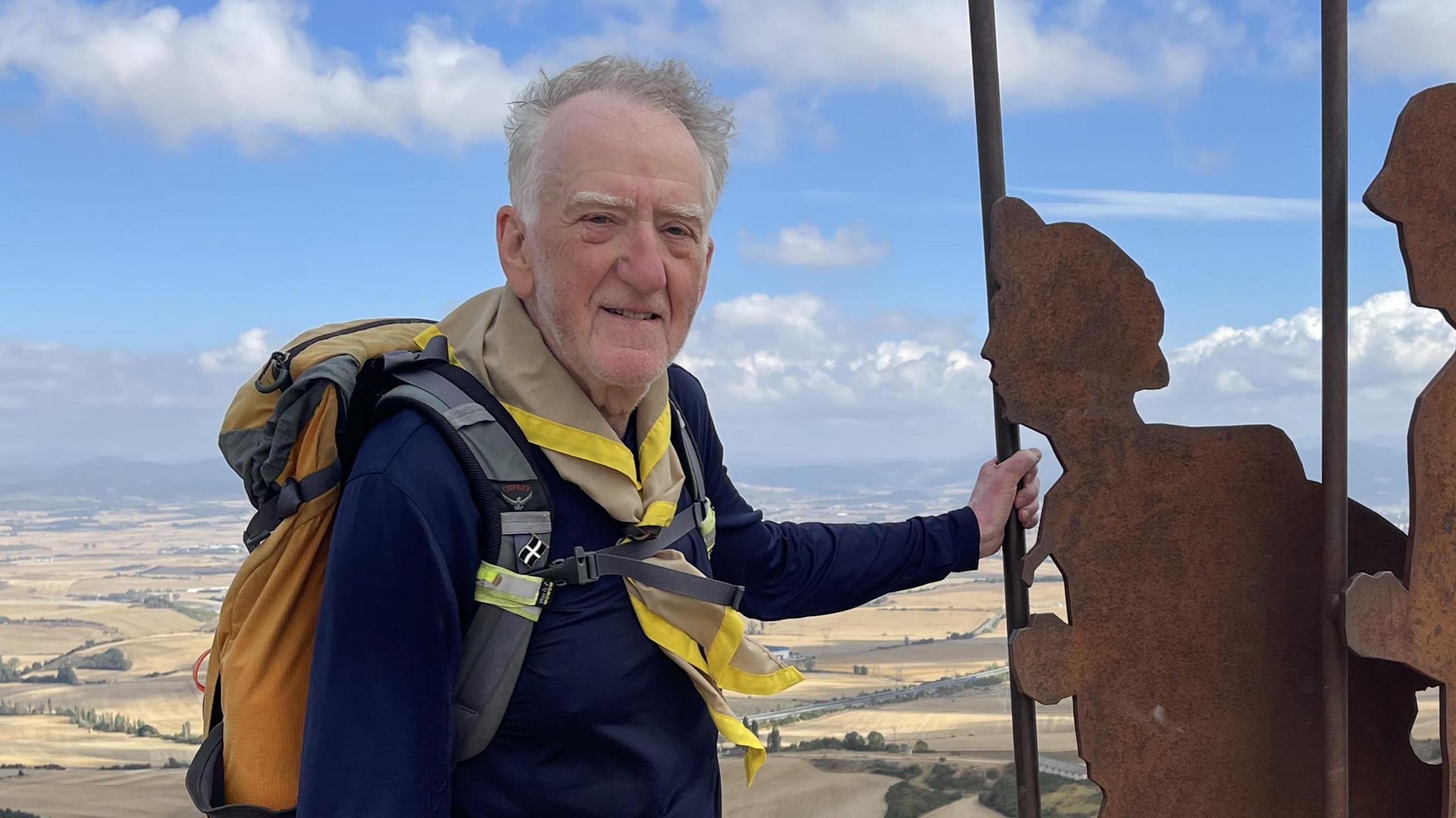David Ensor has short grey hair and grey stubble. He is wearing walking clothing and is standing atop a tor, next to two metal silhouettes of service men and women.