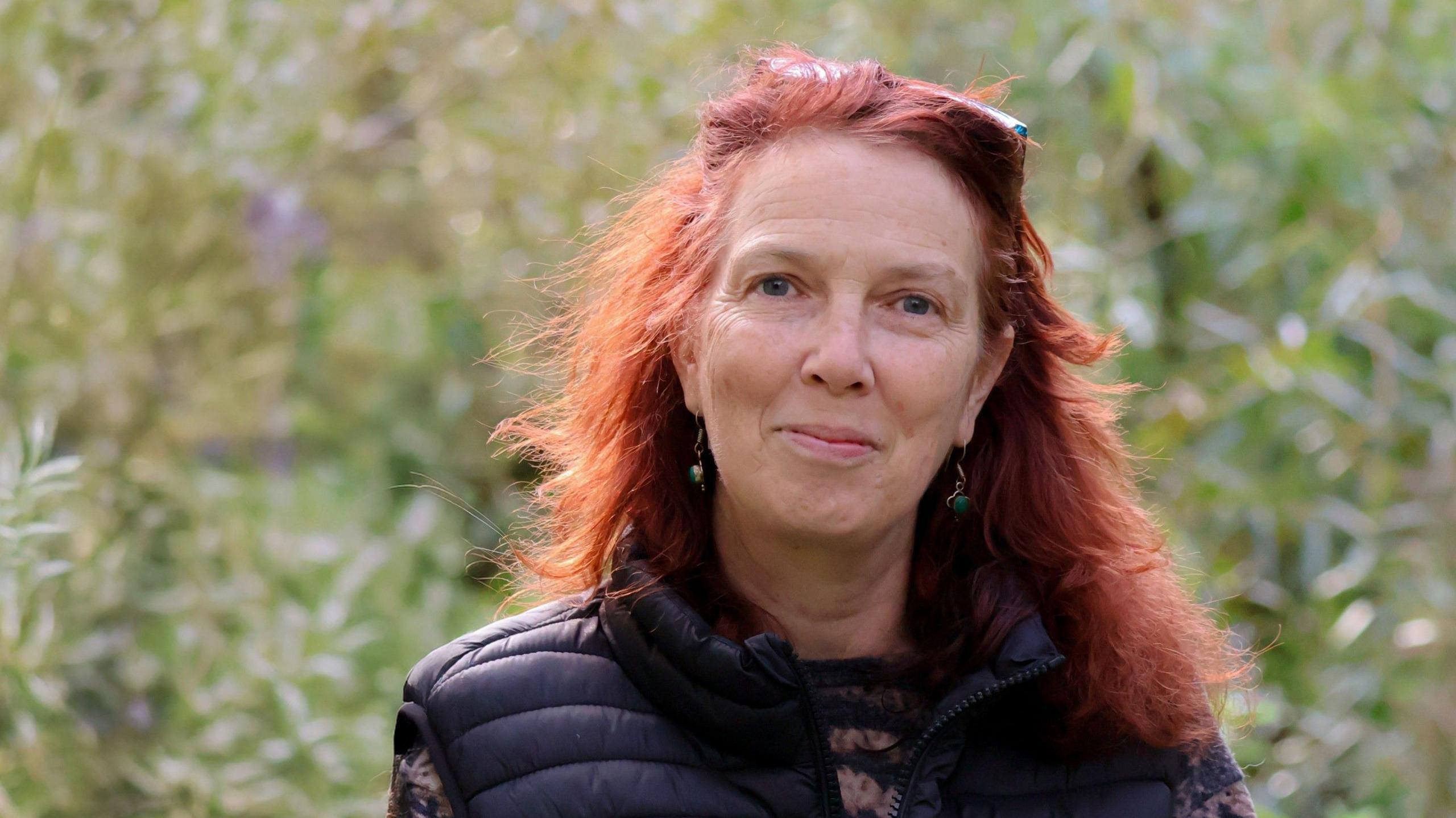 Henri Brocklebank, a woman with long red hair, wears a jersey and a body warmer and stands in a field.