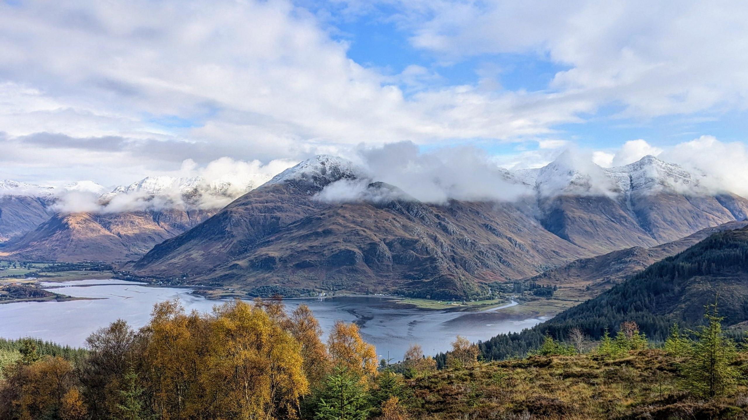 Landscape showing hills from a distance with clouds lingering at the top and a loch at the bottom