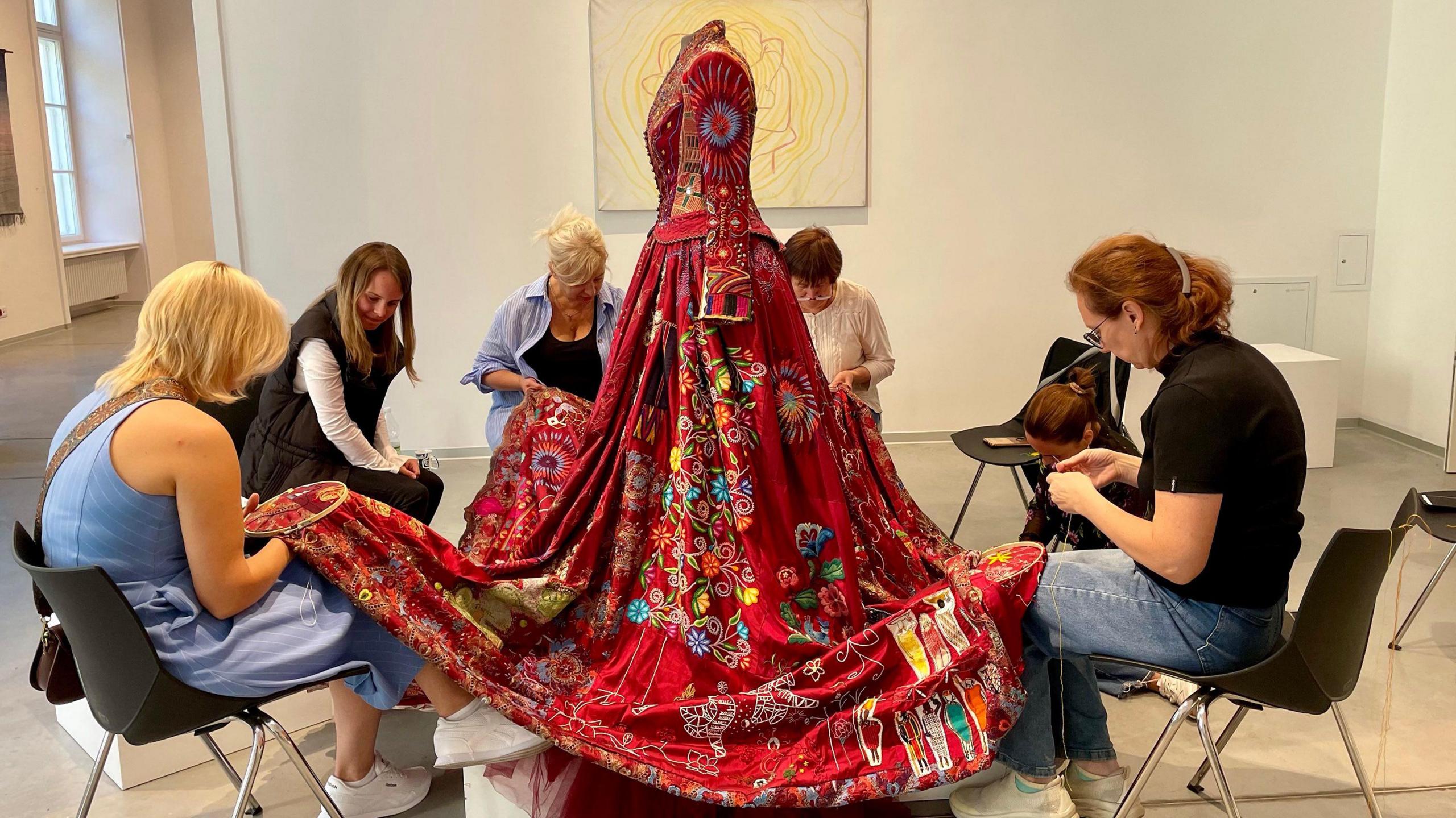 A red dress on a mannequin with women sat on chairs around the dress and embroidering it.