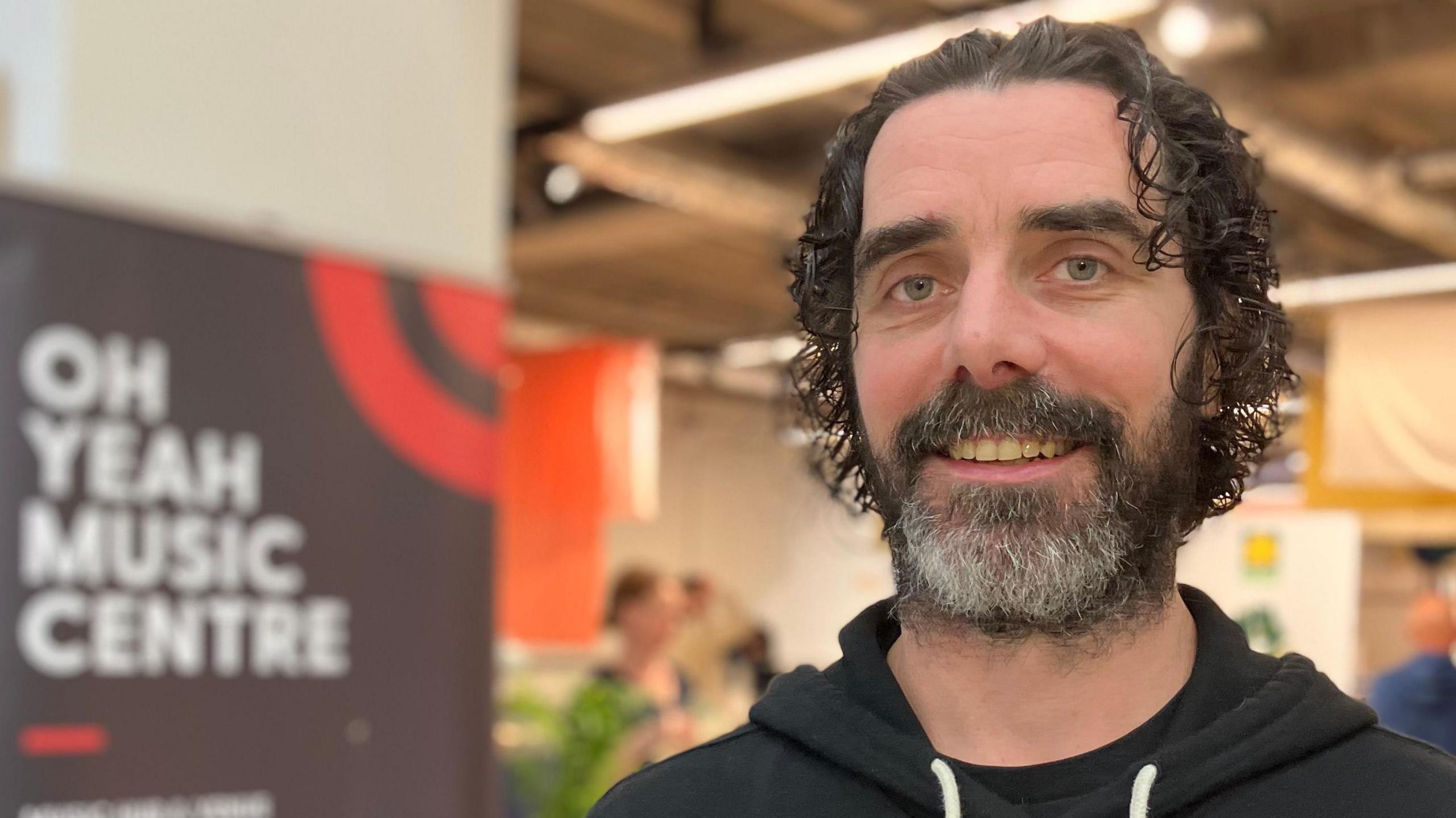 Damien McAdams has curly brown hair and and a beard and moustache. He's wearing a black hoodie and standing in front of a sign saying Oh Yeah Music Centre.