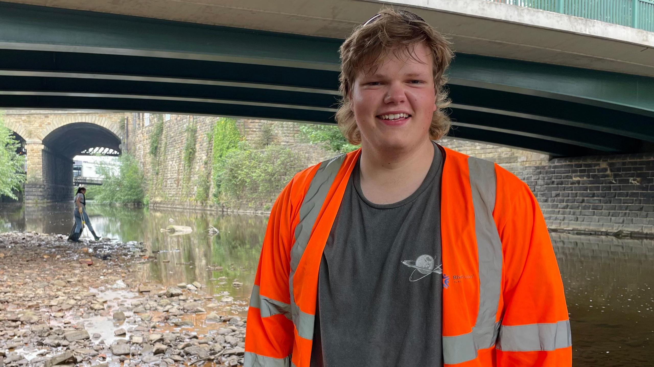 A young man with long blonde hair, wearing a grey T-shirt with a hi-vis jacket, smiles for the camera while standing on a river bank.