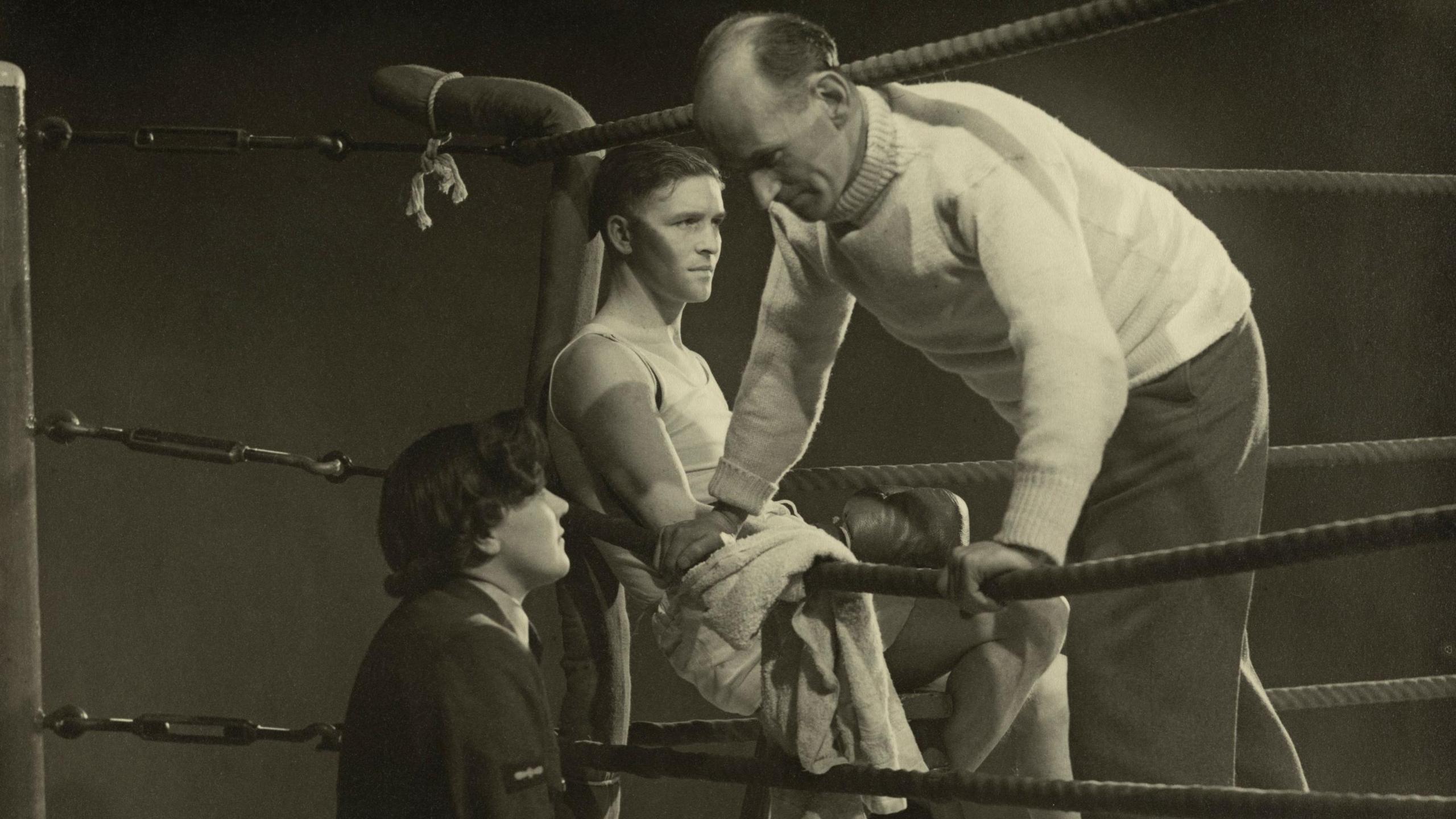 A man leans over the ropes of a boxing ring and looks down at someone in what looks like military uniform. In the boxing ring, a young man is sitting on a chair in the corner, wearing boxing gloves.