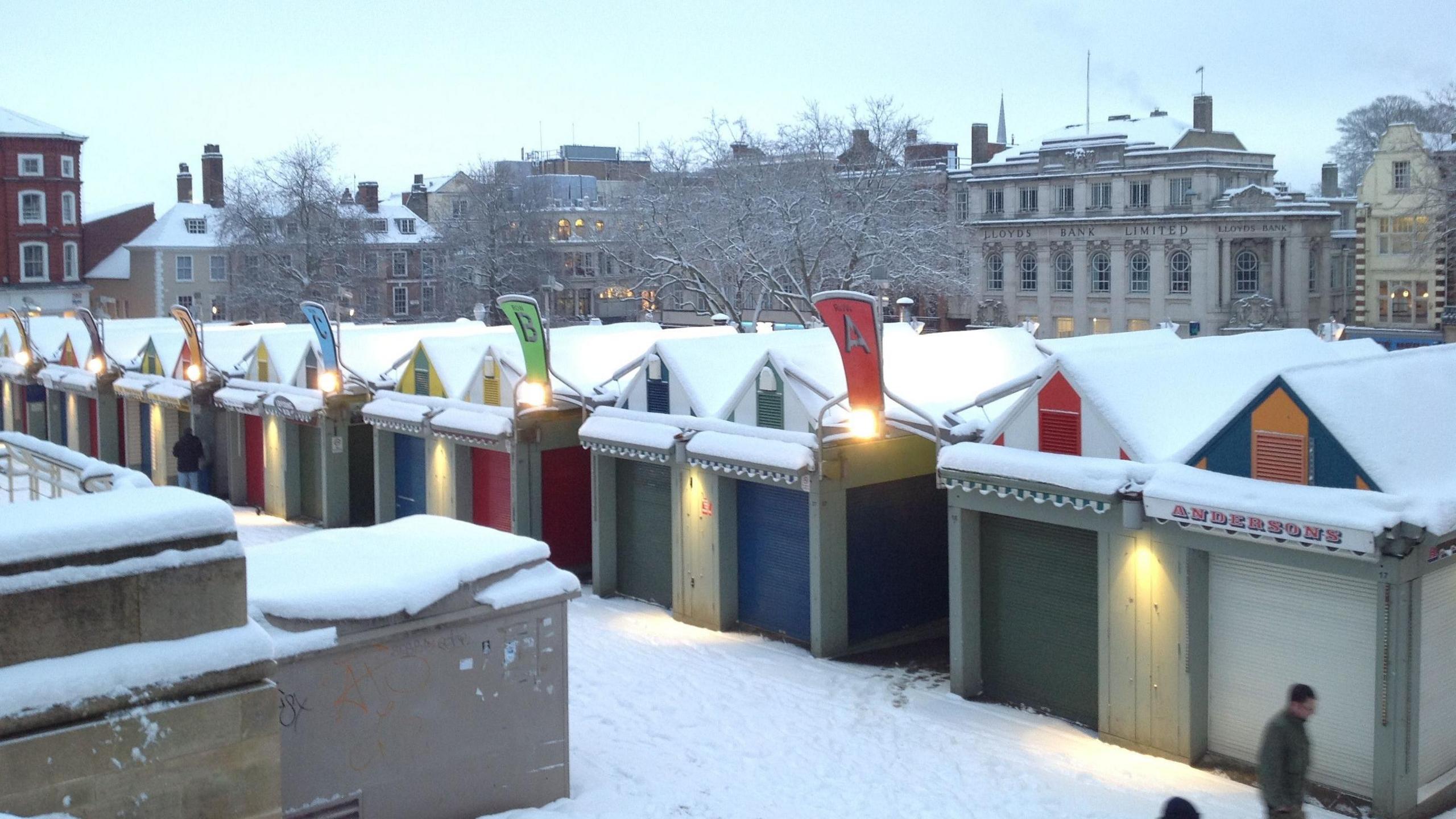 Norwich Market stalls in the snow. They have colourful shutters and historic city centre buildings can be seen behind them. These include Jarrolds department store.