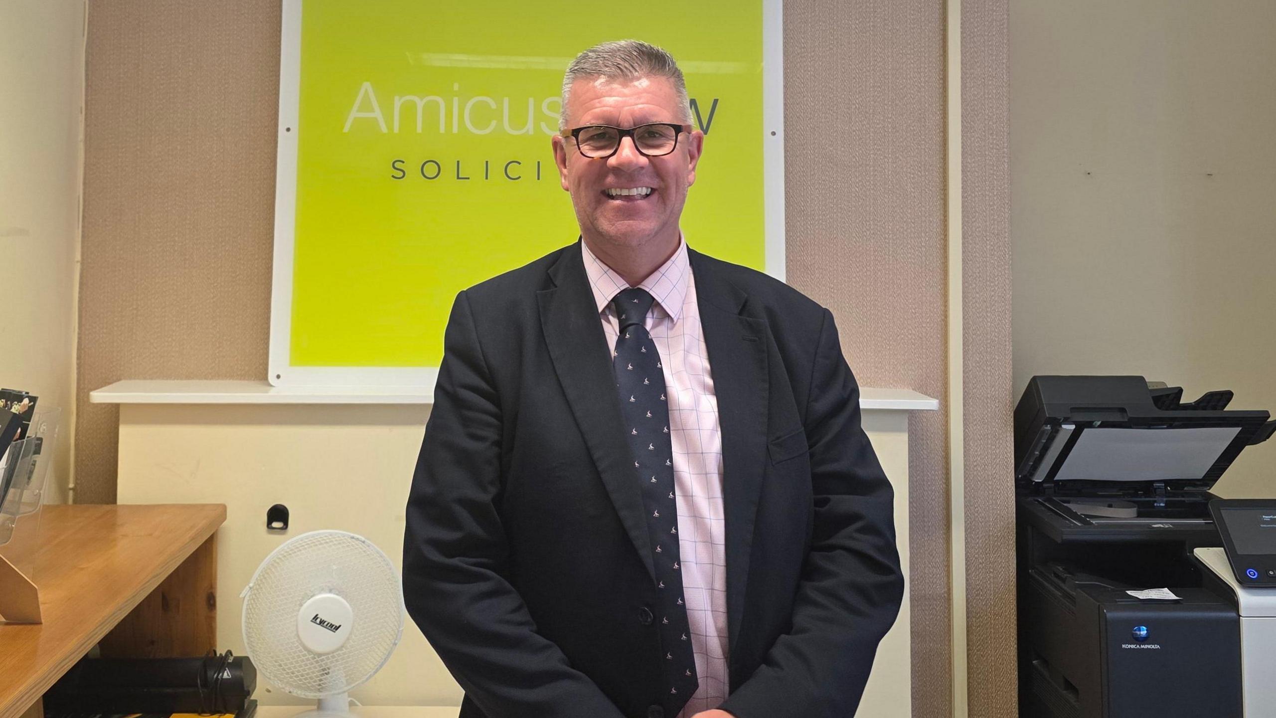 A man in a suit is standing in an office with both hands folded in front of him. He is wearing black glasses and smiling at the camera.