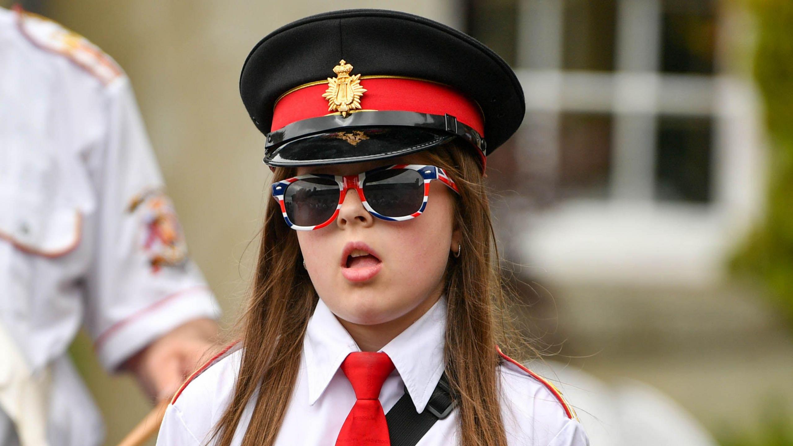 A young girl wears a pair of sunglasses with the Union Jack flag on them. She is also wearing a white band uniform and a red tie.