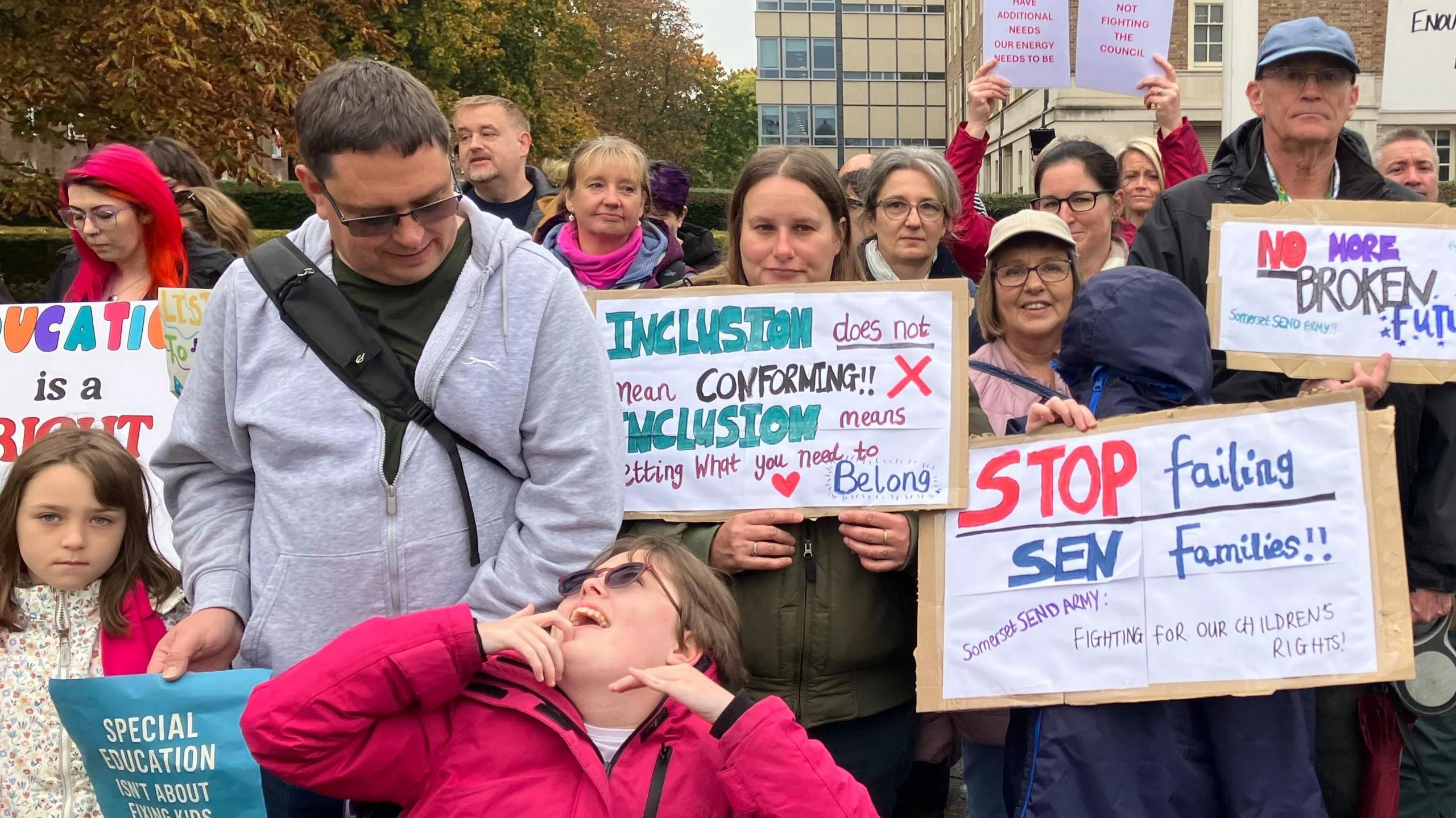 A group of people holding placards reading things like 'stop failing sen families' and a girl in a wheelchair in the foreground looking up at an adult