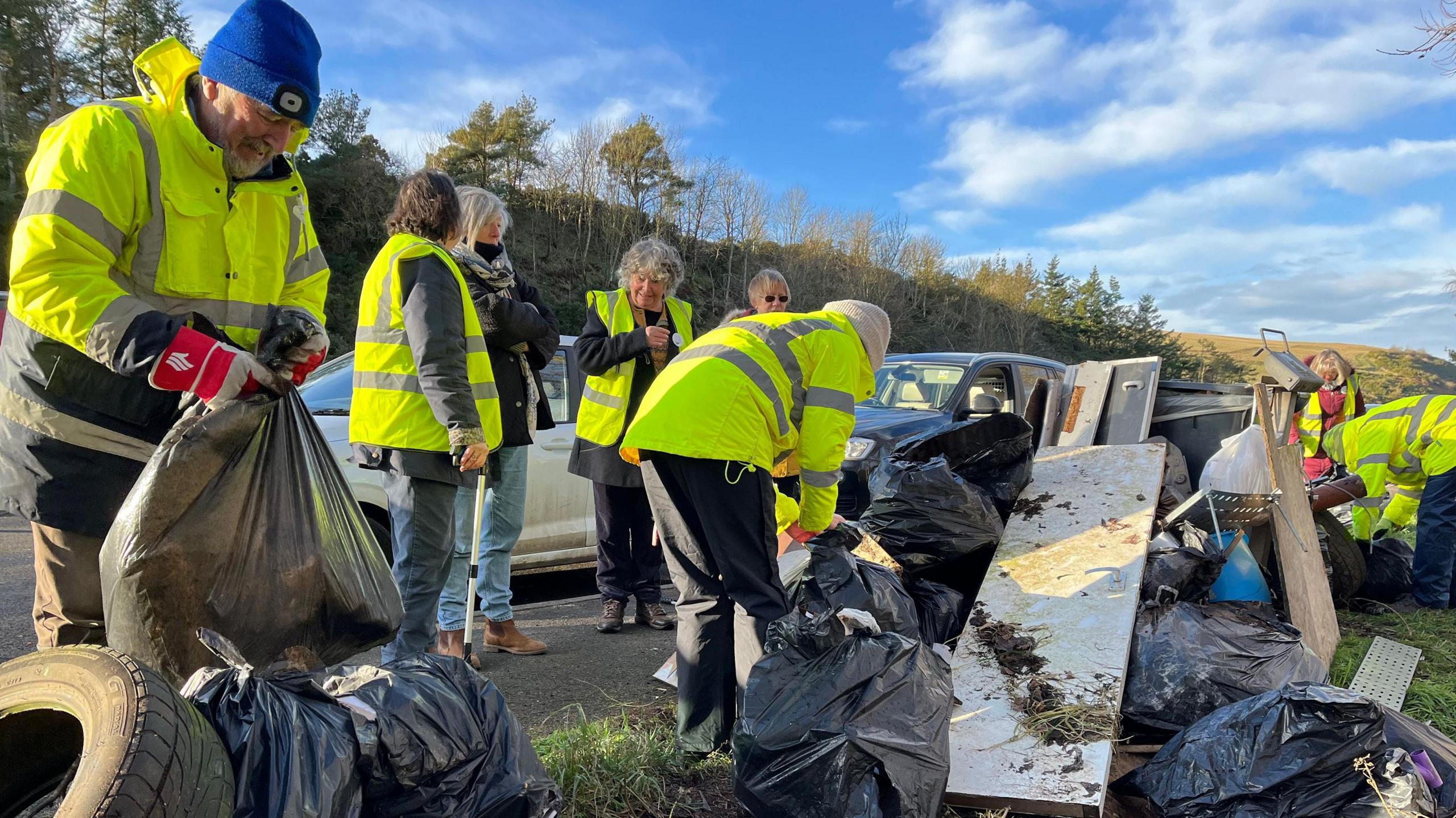 A man in high visibility yellow jacket and blue hat ties a black bin bag, while behind him other people in high visibility jackets collect filled black bags.