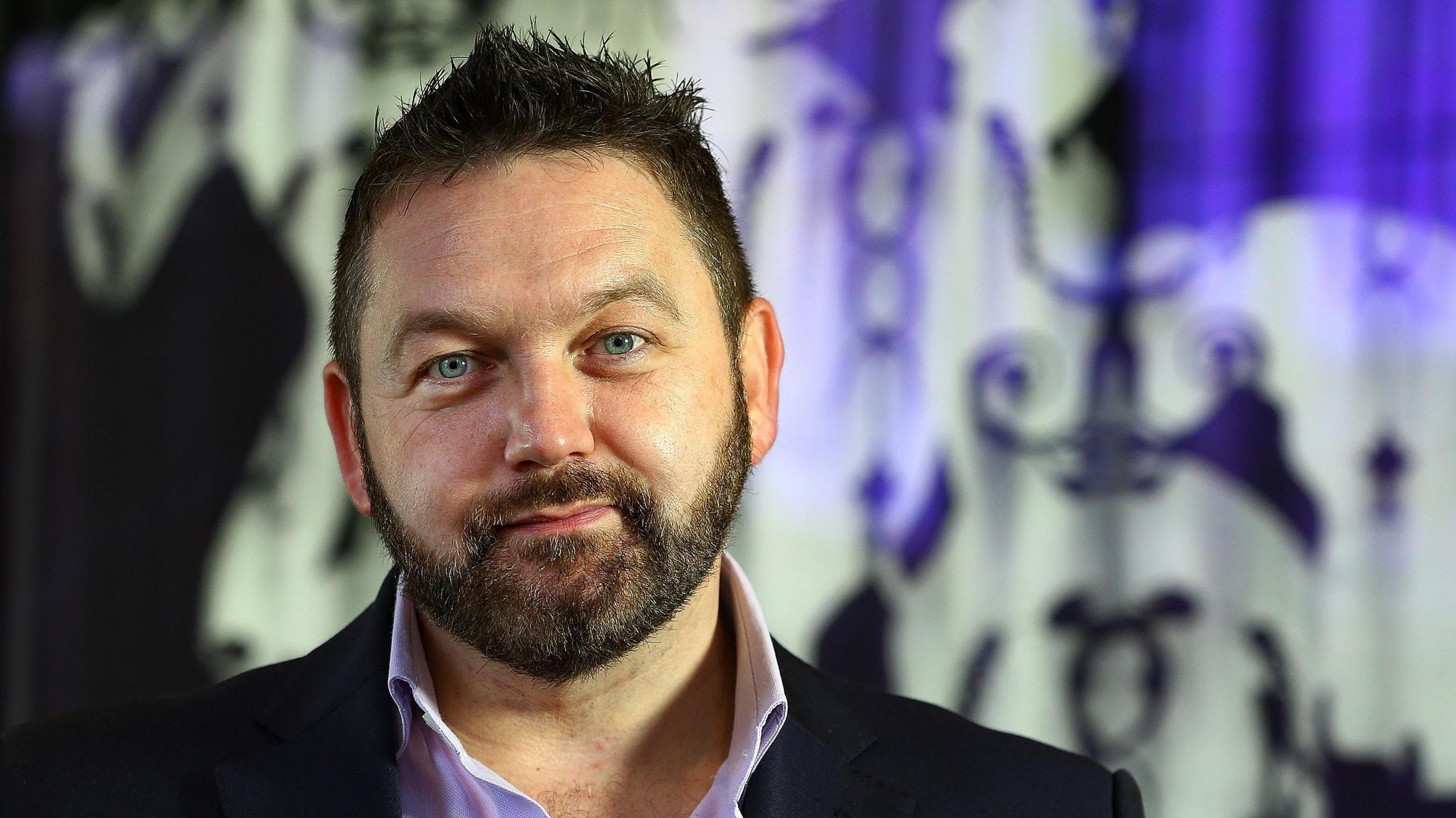 William Crawley, a man with dark hair and a short beard, wearing a lilac shirt and dark jacket