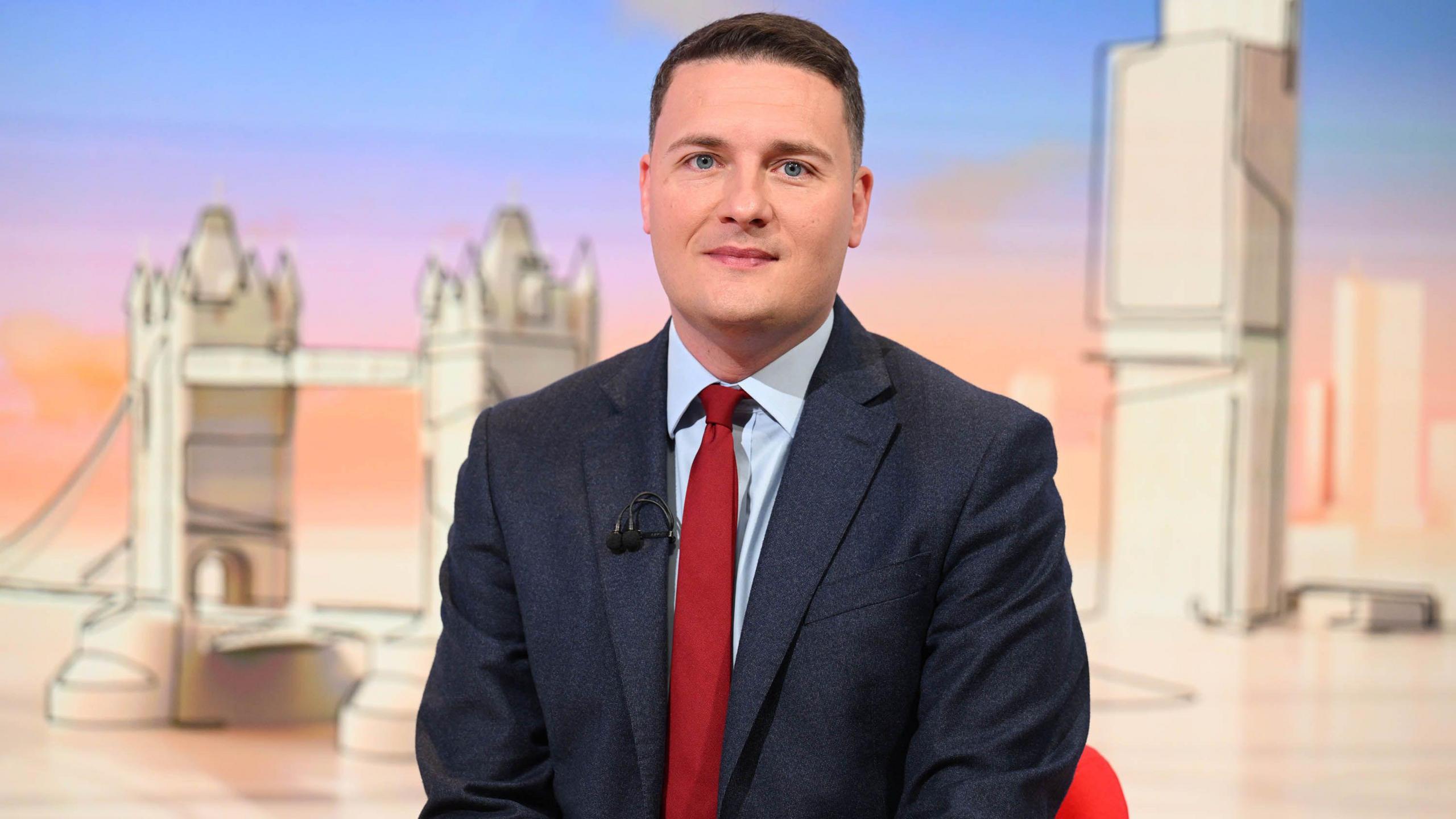 Mid shot of Health Secretary Wes Streeting sitting down in a red chair in some kind of studio with an artistic line drawing representation of Tower Bridge and some other buildings in the background. Mr Streeting is wearing a navy suit with a red tie and light blue shirt.